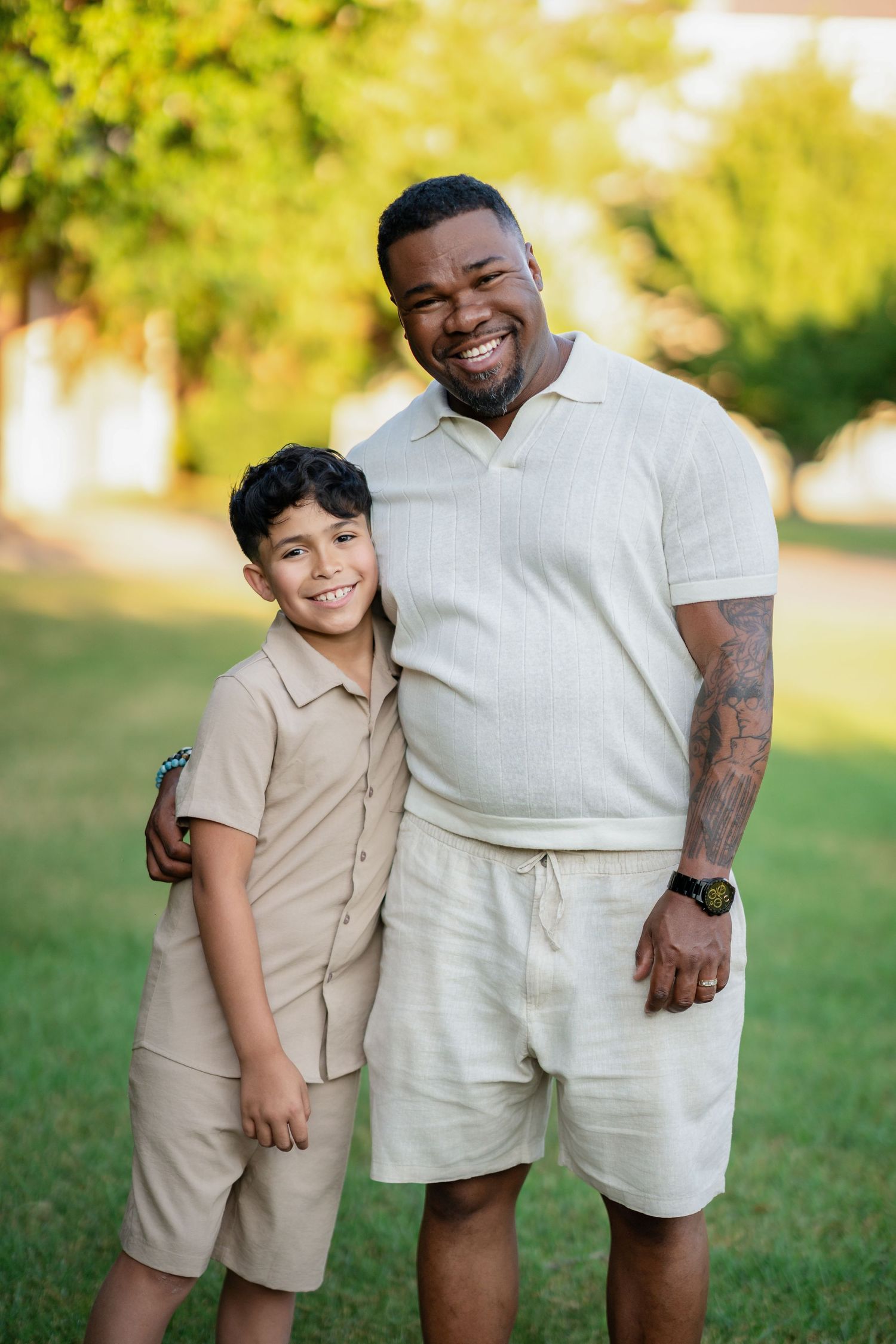 A smiling man with tattoos and a young boy pose together outdoors on green grass in matching beige outfits.