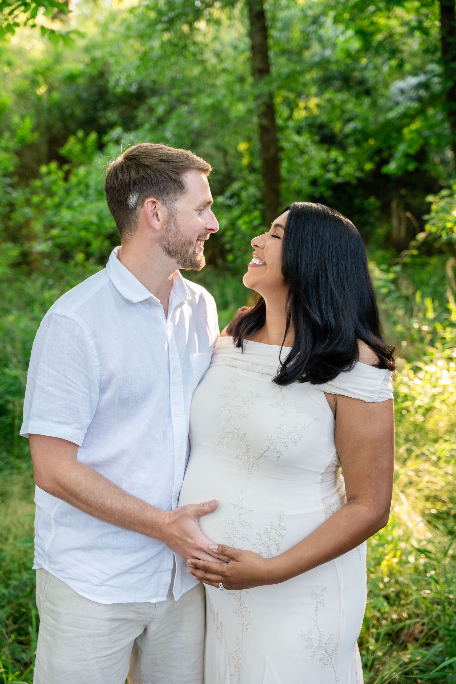 Smiling expectant couple embracing in a sunlit green forest, the man in a white shirt holding the pregnant woman's belly in a white dress.