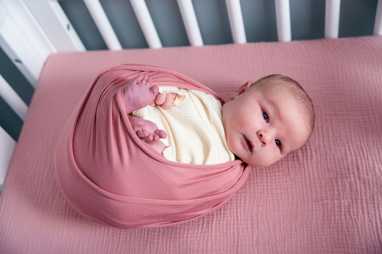 Newborn baby swaddled in pink wrap lying in a white crib on a dusty rose muslin blanket.
