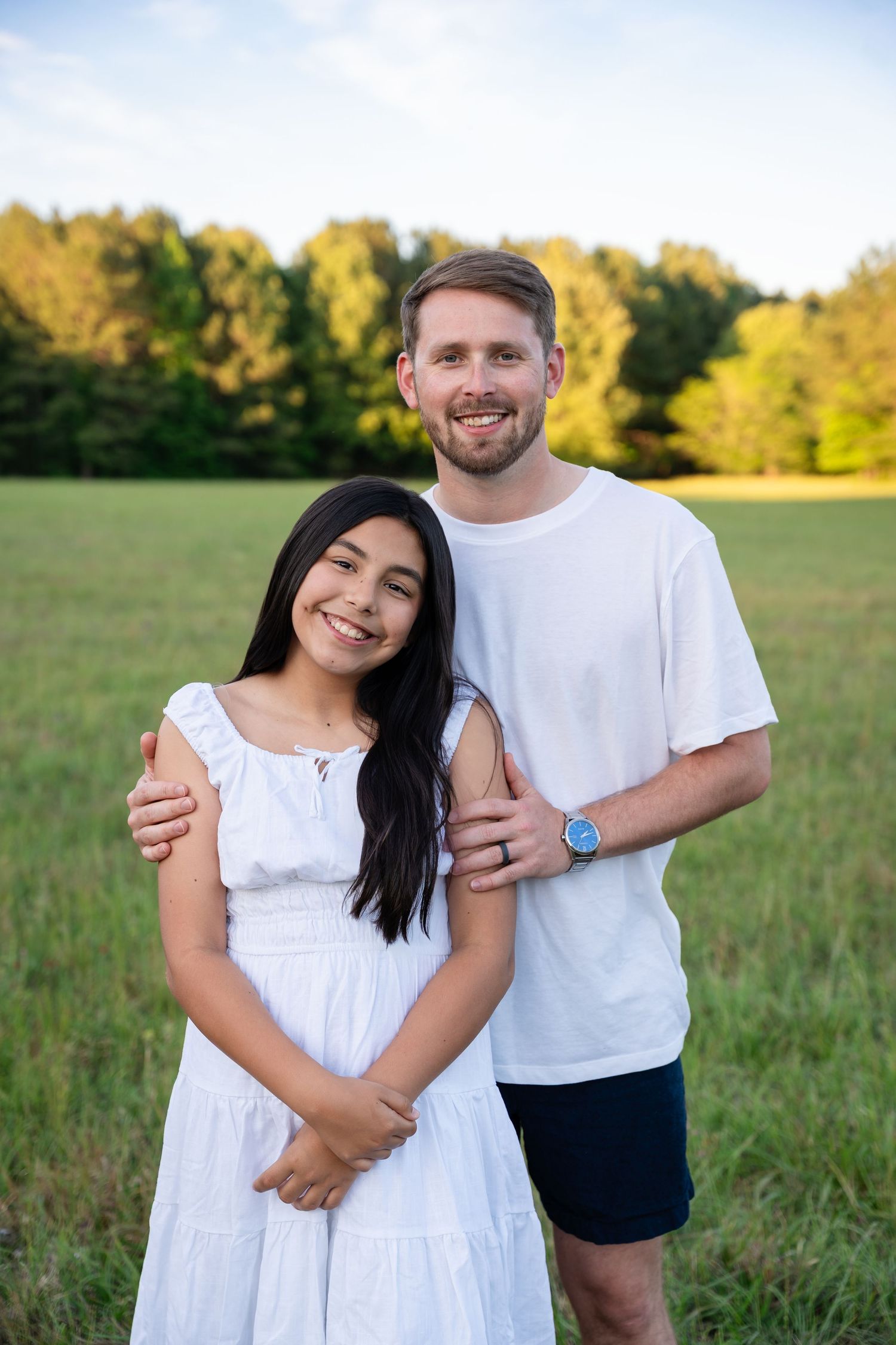 Smiling couple in white outfits posing in a green field, man embracing woman with trees in the background.