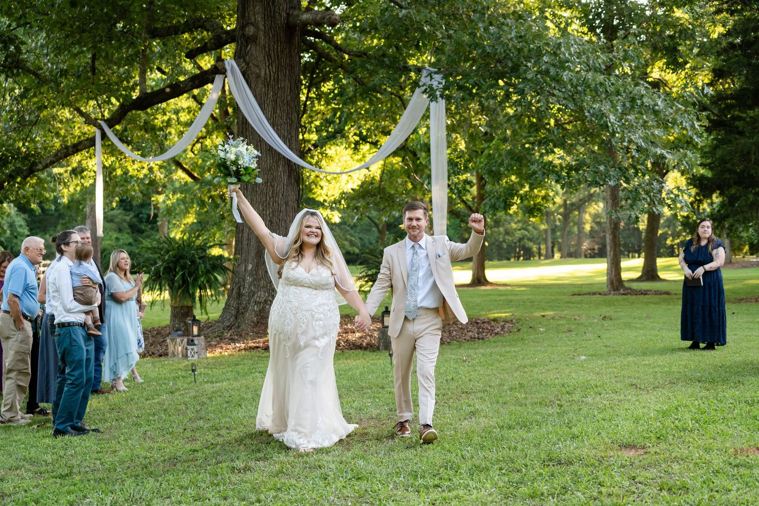 Joyful newlyweds celebrate under floral arch on green lawn at outdoor wedding ceremony with guests watching.