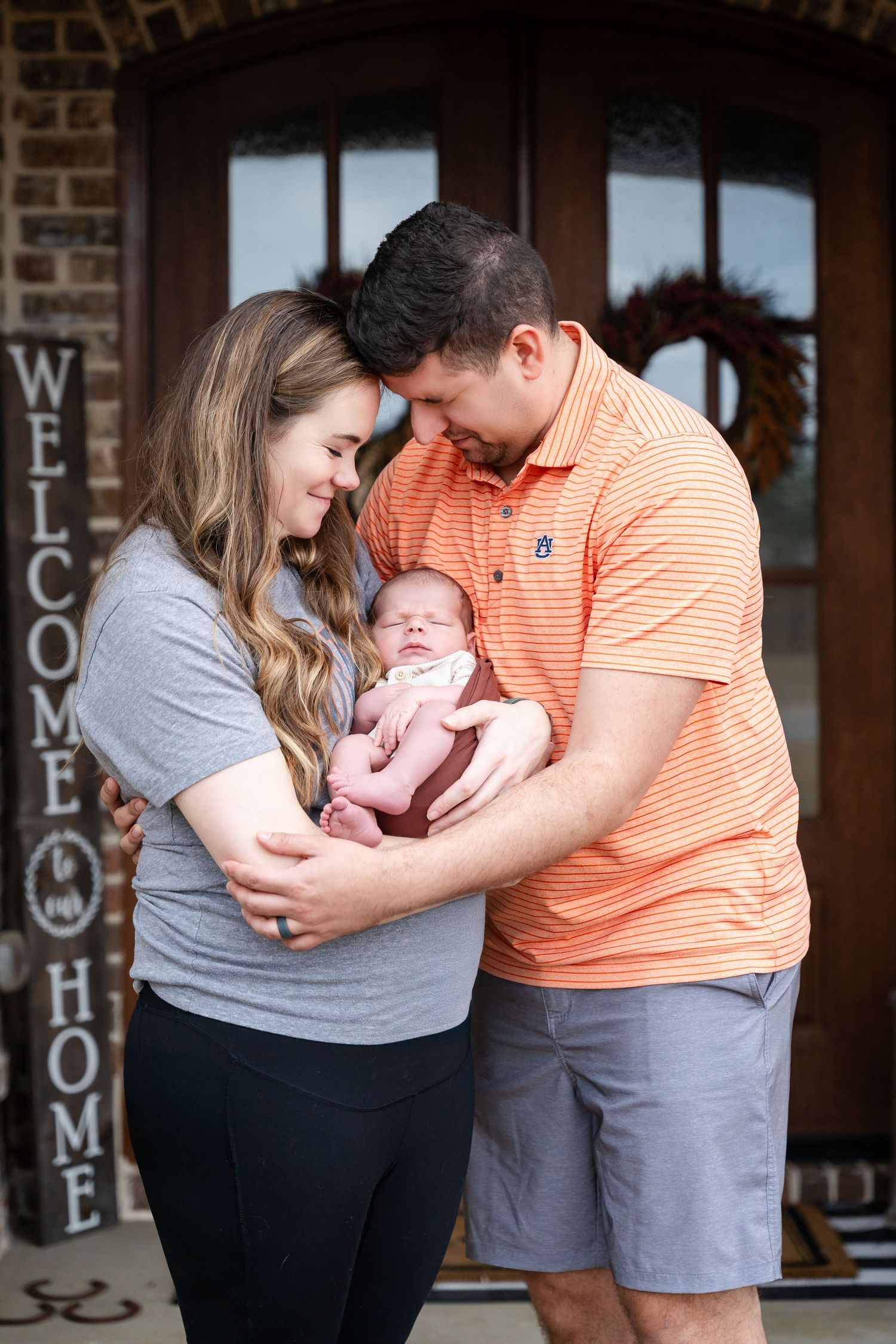 A tender family moment as parents cradle their newborn baby in front of a welcome sign at their home entrance.