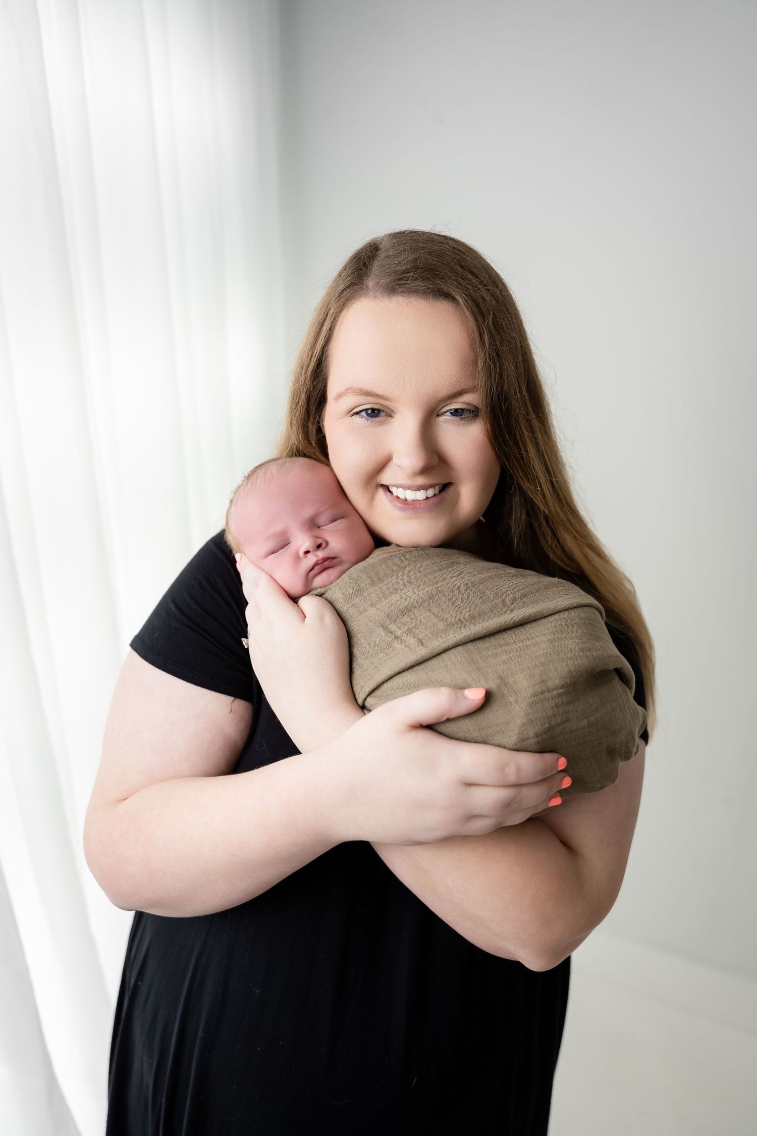 Mother in black dress holding wrapped baby against white curtain background.