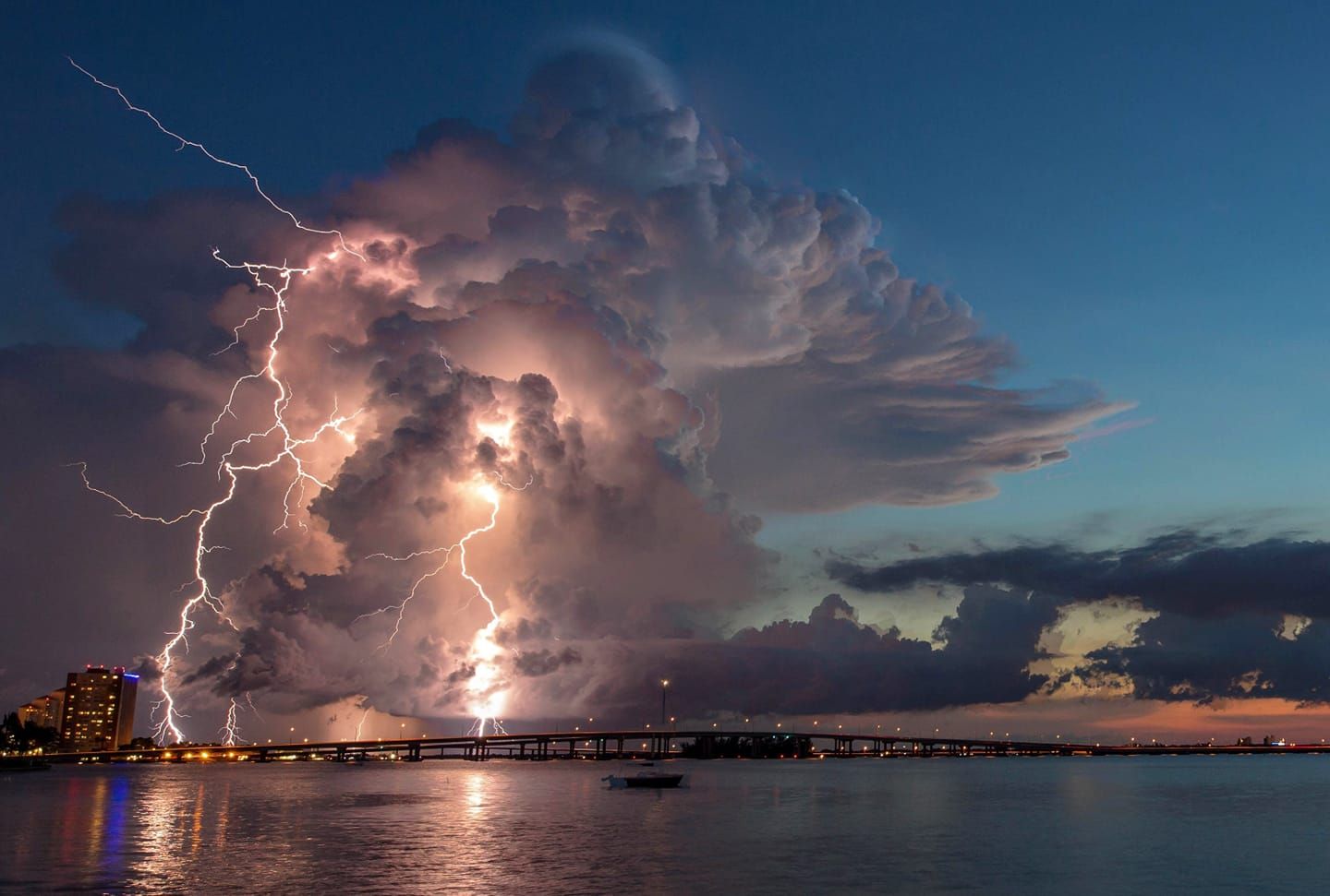 Lightning strikes illuminate a dramatic storm cloud over a coastal pier at sunset with city lights glowing in the distance.