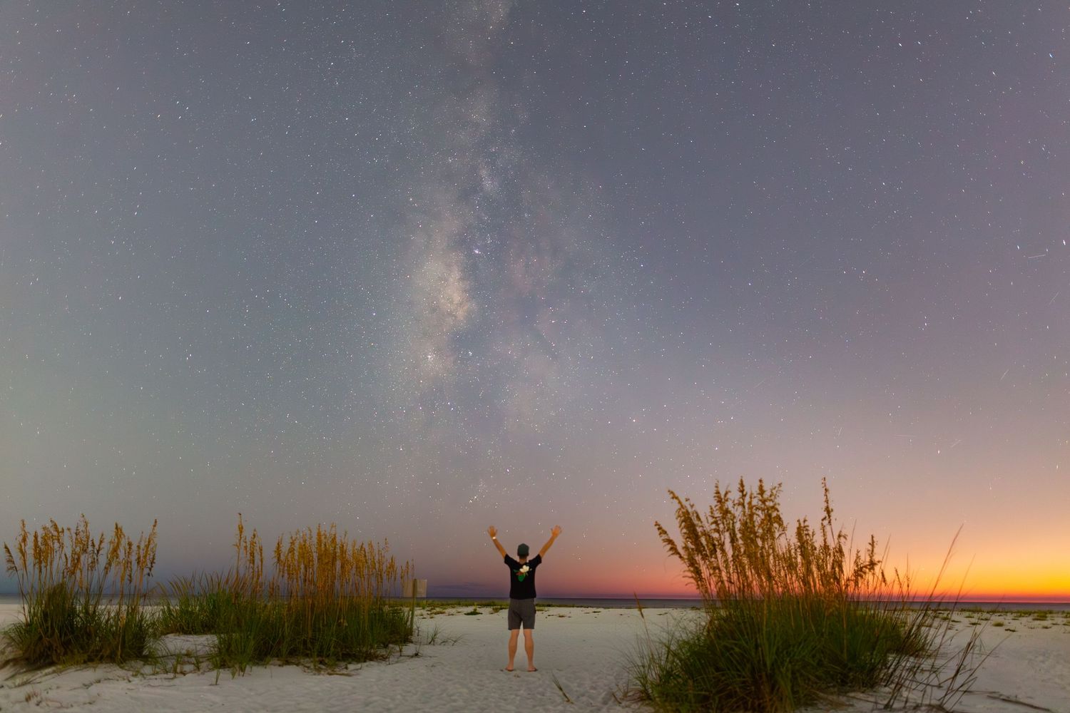 Silhouette of a person standing with raised arms against a colorful sunset sky over grassy wetlands.