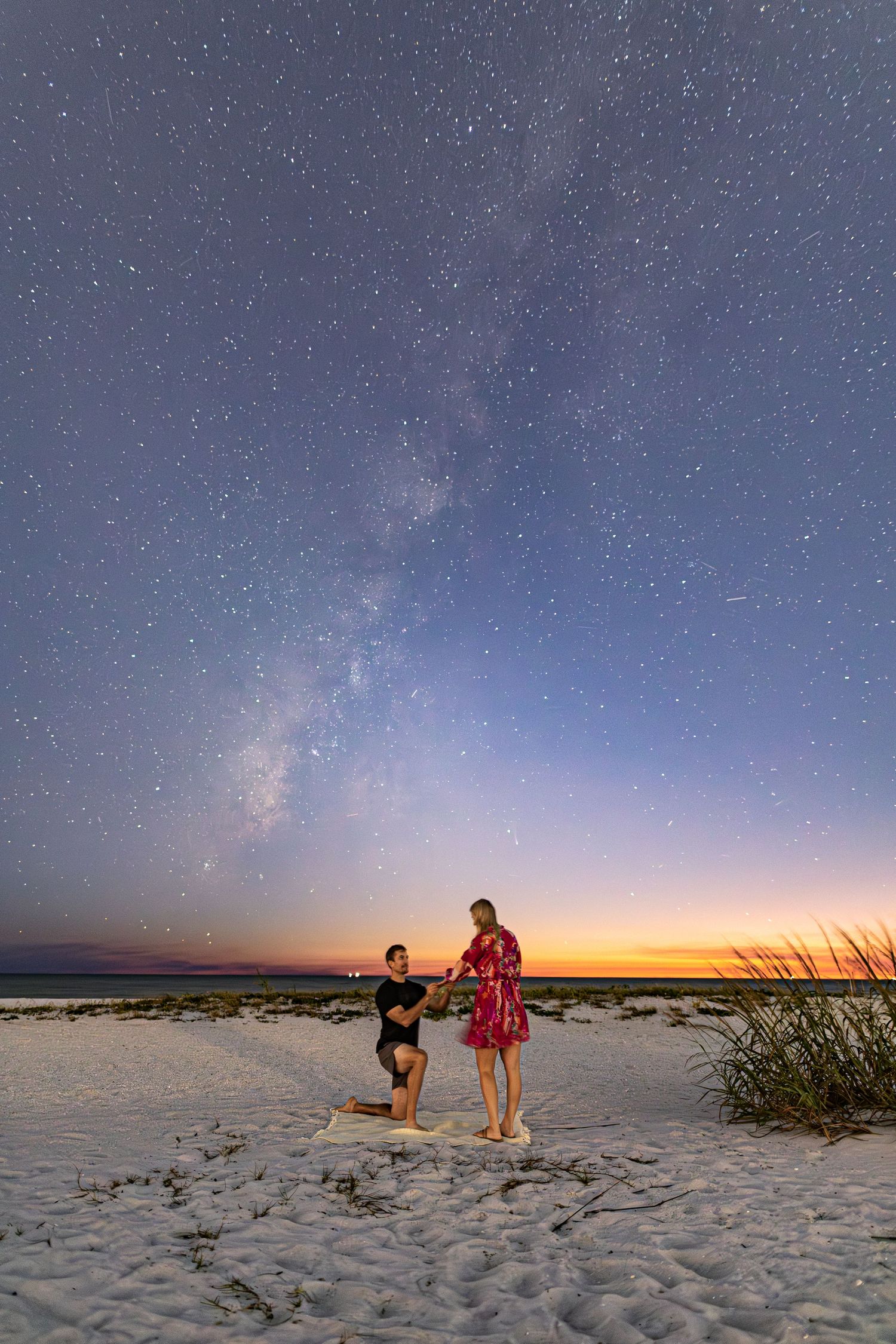 Two people stand on a beach at sunset with the Milky Way galaxy visible in the starry night sky above.