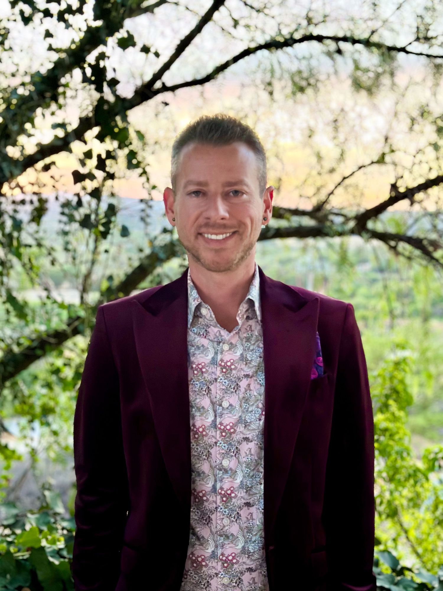 A person wearing a burgundy blazer and floral shirt poses outdoors with blooming trees in the background.