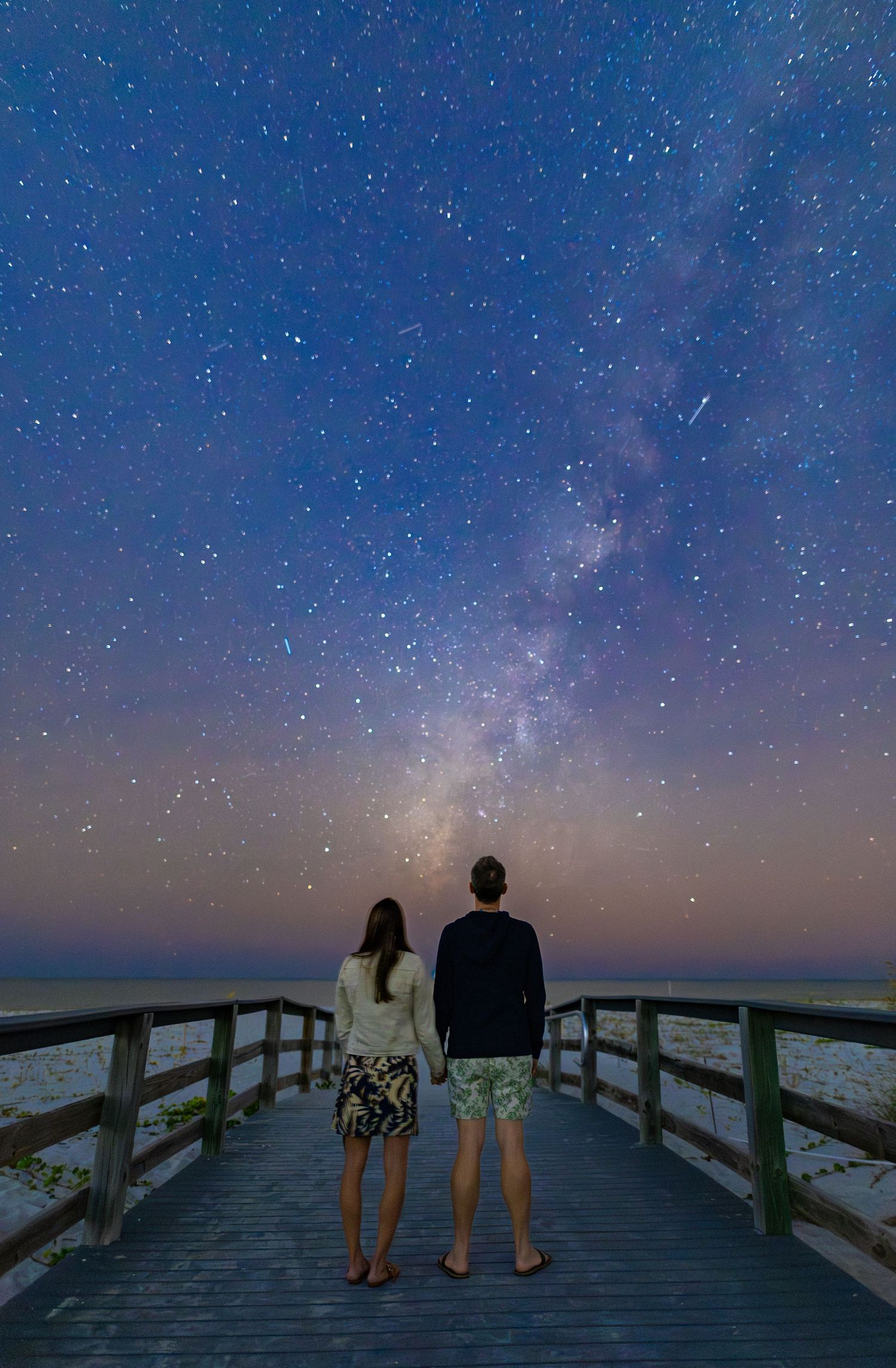 People stand on a wooden boardwalk under a starlit sky with the Milky Way galaxy visible above.