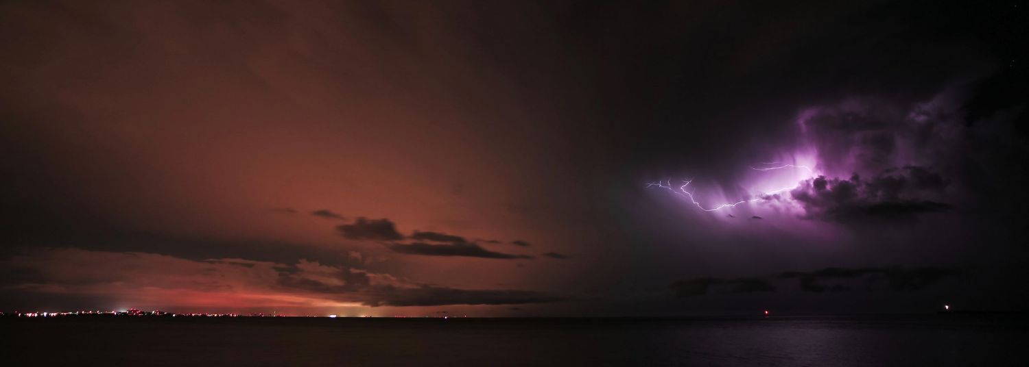 Lightning illuminates a dramatic stormy sky over a dark landscape at night with purple and orange hues.