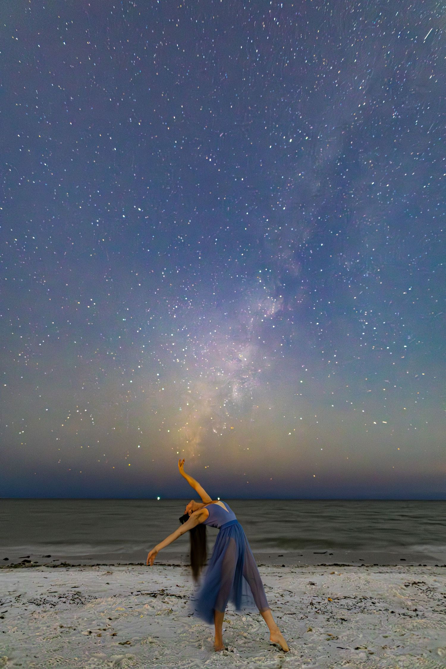 A person photographs the starry night sky on a beach with the Milky Way visible in the dark blue horizon.