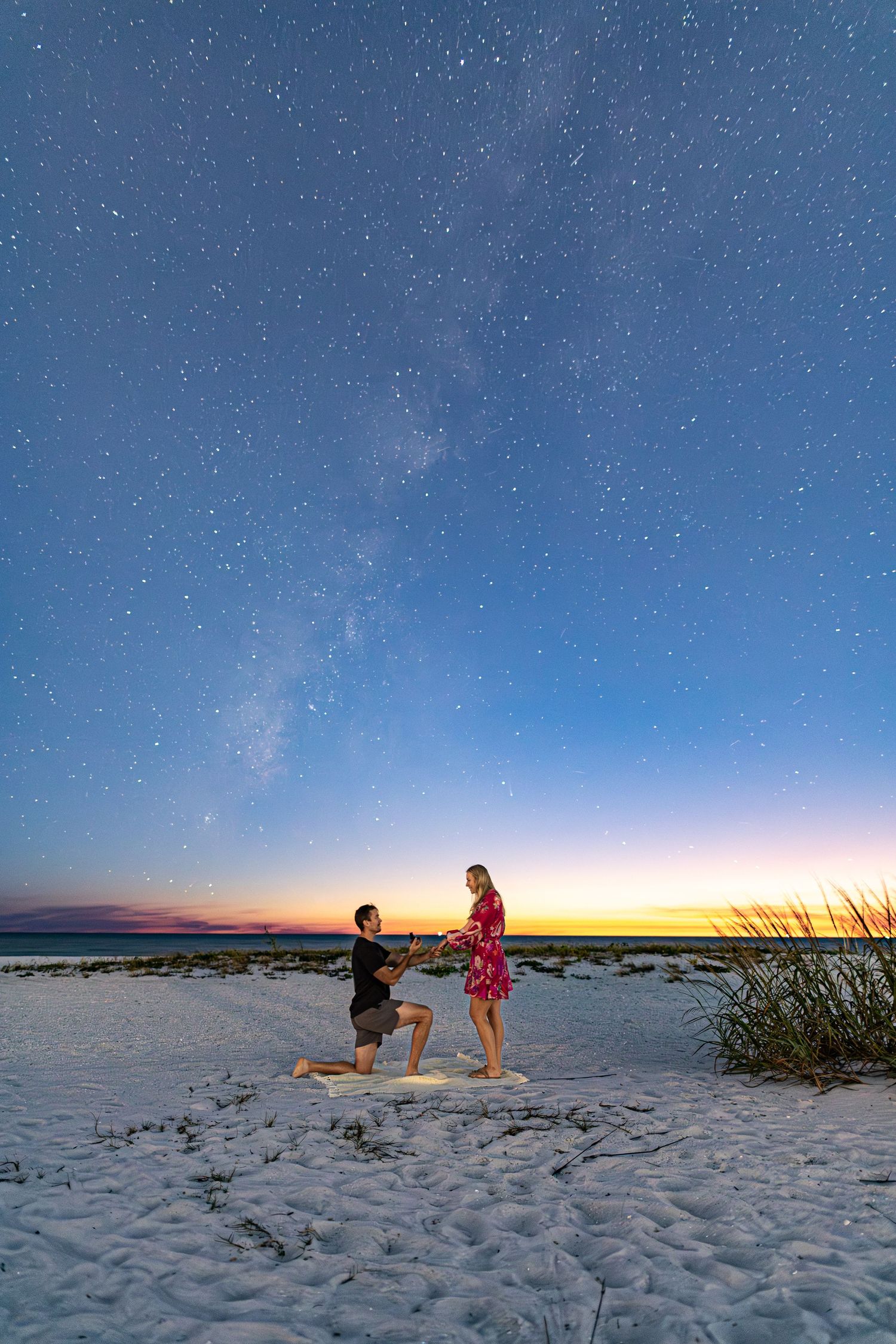 A romantic beach proposal scene at sunset with two figures silhouetted against a colorful sky over white sand.