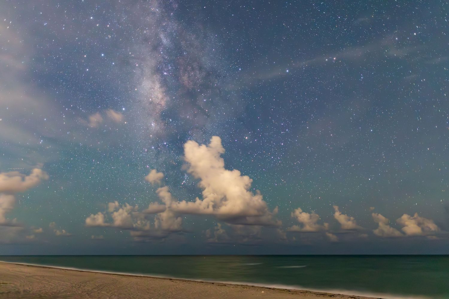 Starry night sky with fluffy clouds over turquoise ocean waters and sandy beach under moonlit conditions.