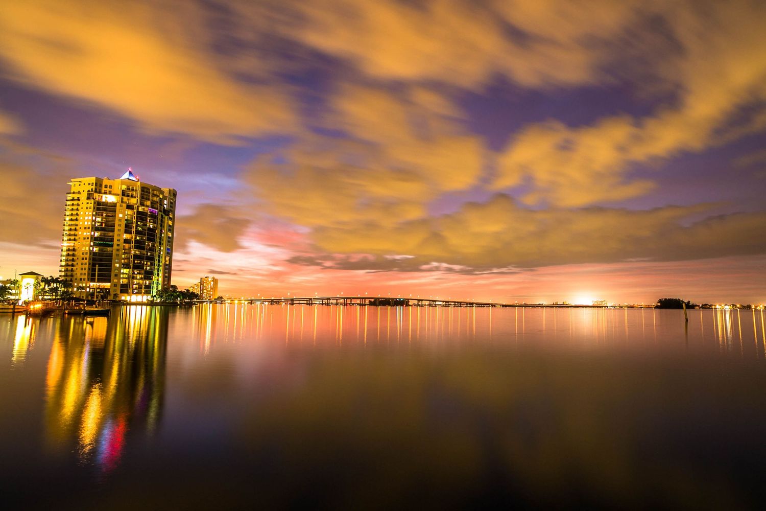 Dramatic orange sunset over calm waters with city skyline silhouettes and illuminated buildings reflecting on the bay.