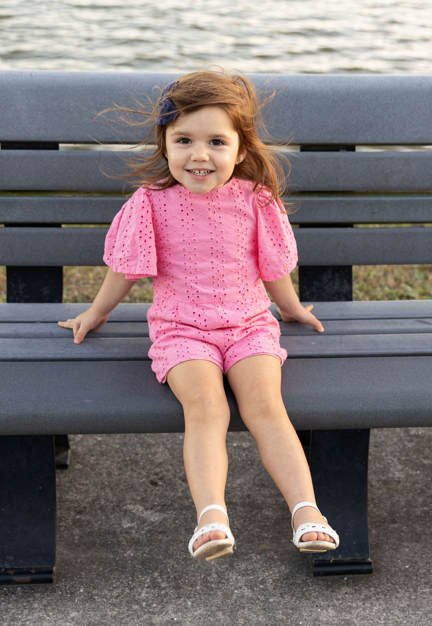 A child in a pink lace outfit sits on a dark bench with their legs dangling, showing white sandals.