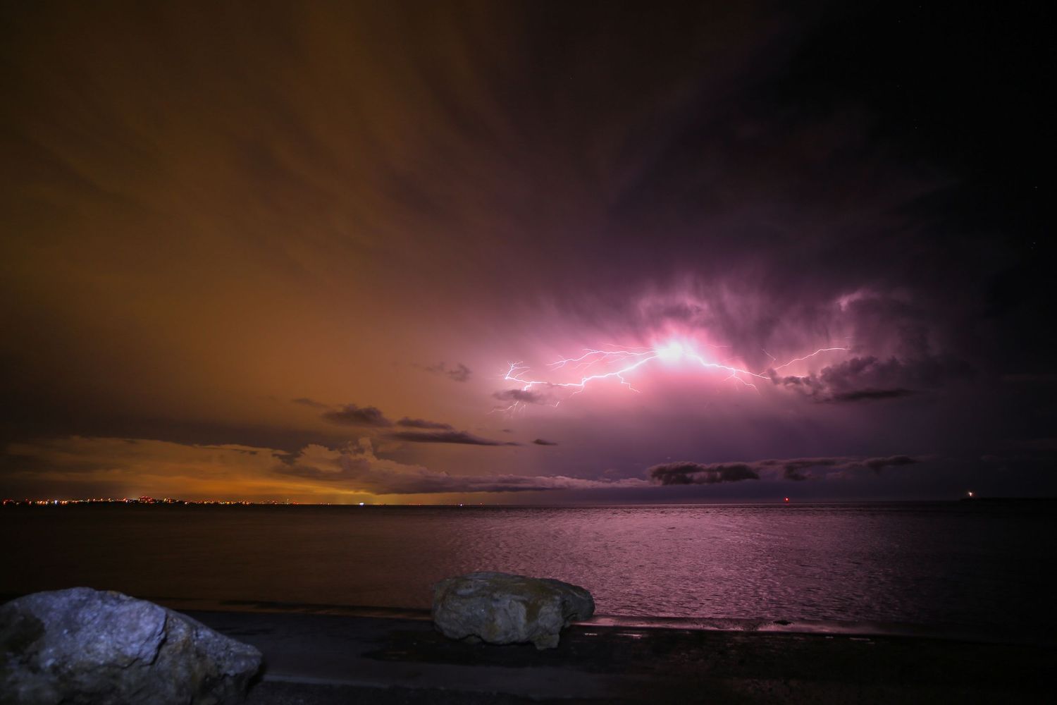 Lightning illuminates a dramatic purple and pink stormy sky over a dark coastline at night.