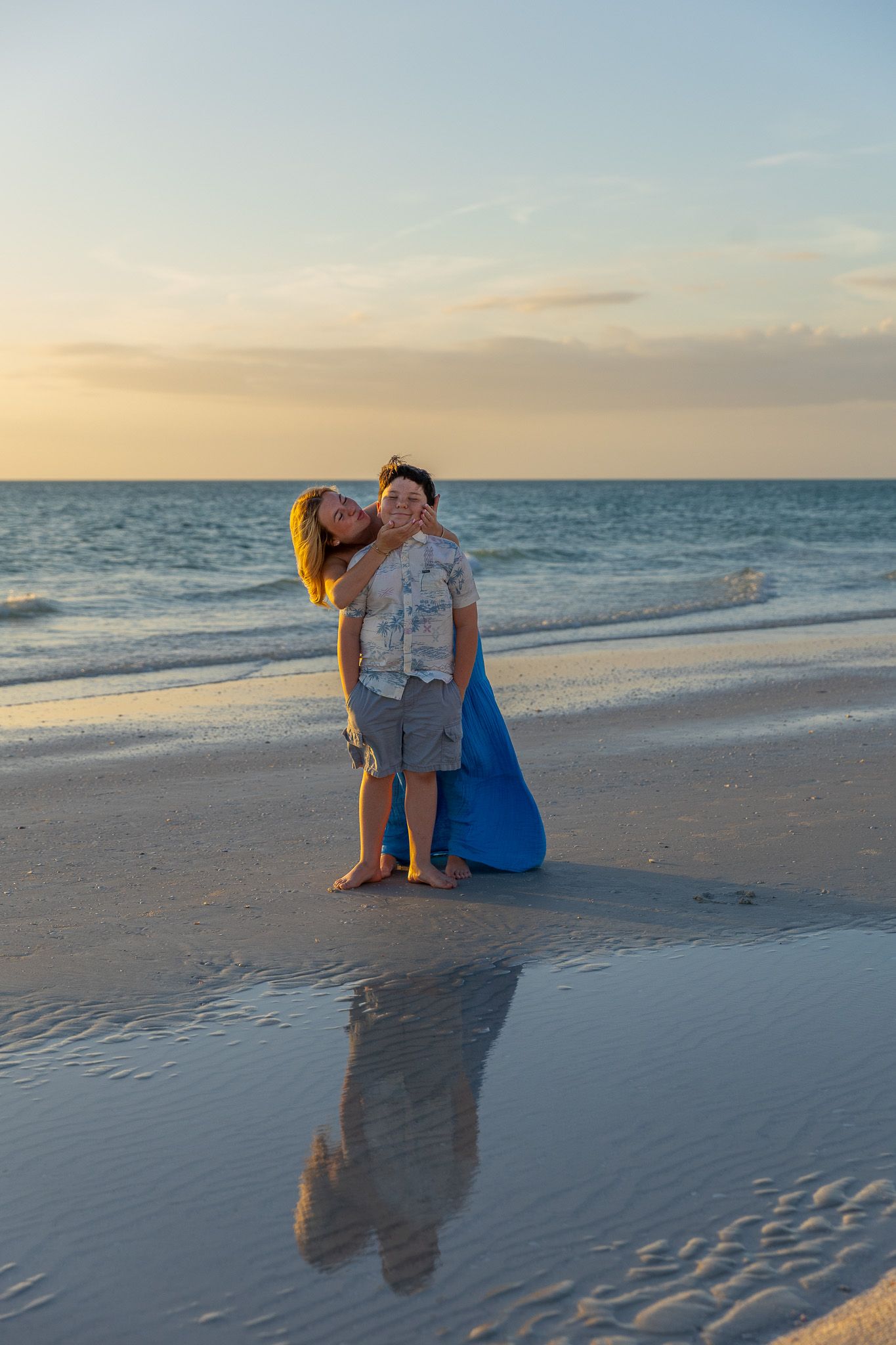 A couple embraces on a peaceful beach at sunset, their reflection visible in the wet sand along the shoreline.
