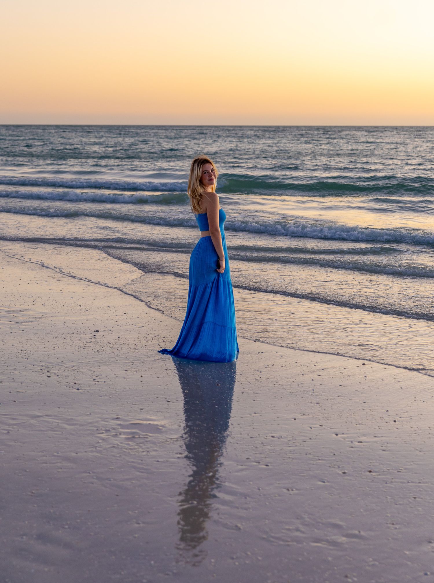 A figure in a flowing blue dress stands at the edge of the ocean during a serene sunset at the beach.