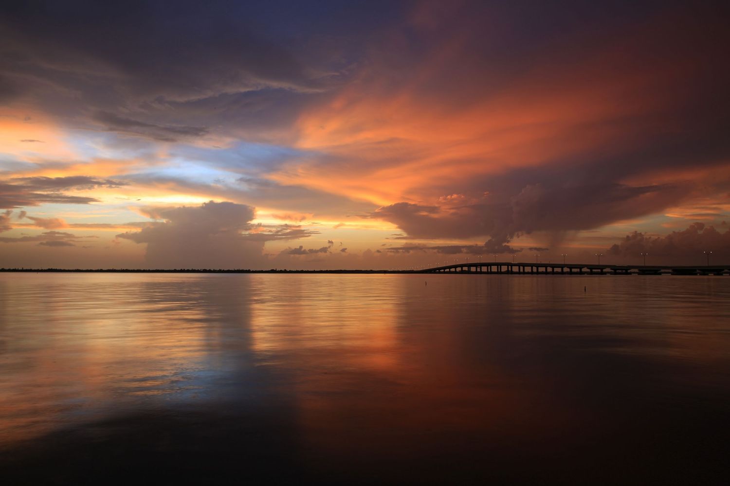 Vibrant orange and purple sunset reflects on calm waters with a distant pier silhouetted against the colorful sky.