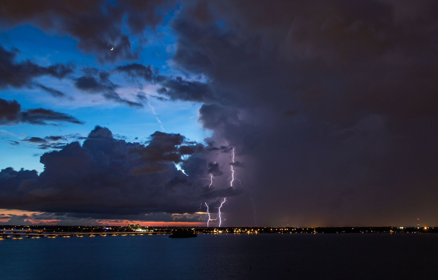 Lightning bolts illuminate dramatic storm clouds over city lights and water at night creating an electrifying display.