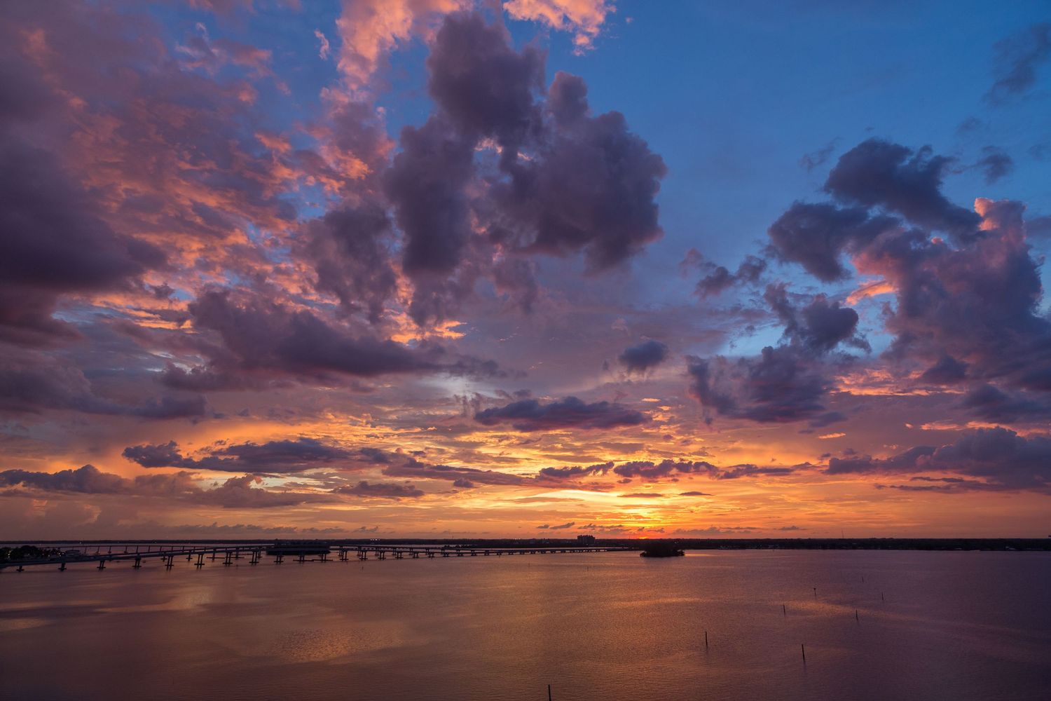 Dramatic sunset sky with vibrant orange clouds reflecting over calm water and a silhouetted pier stretching to horizon.