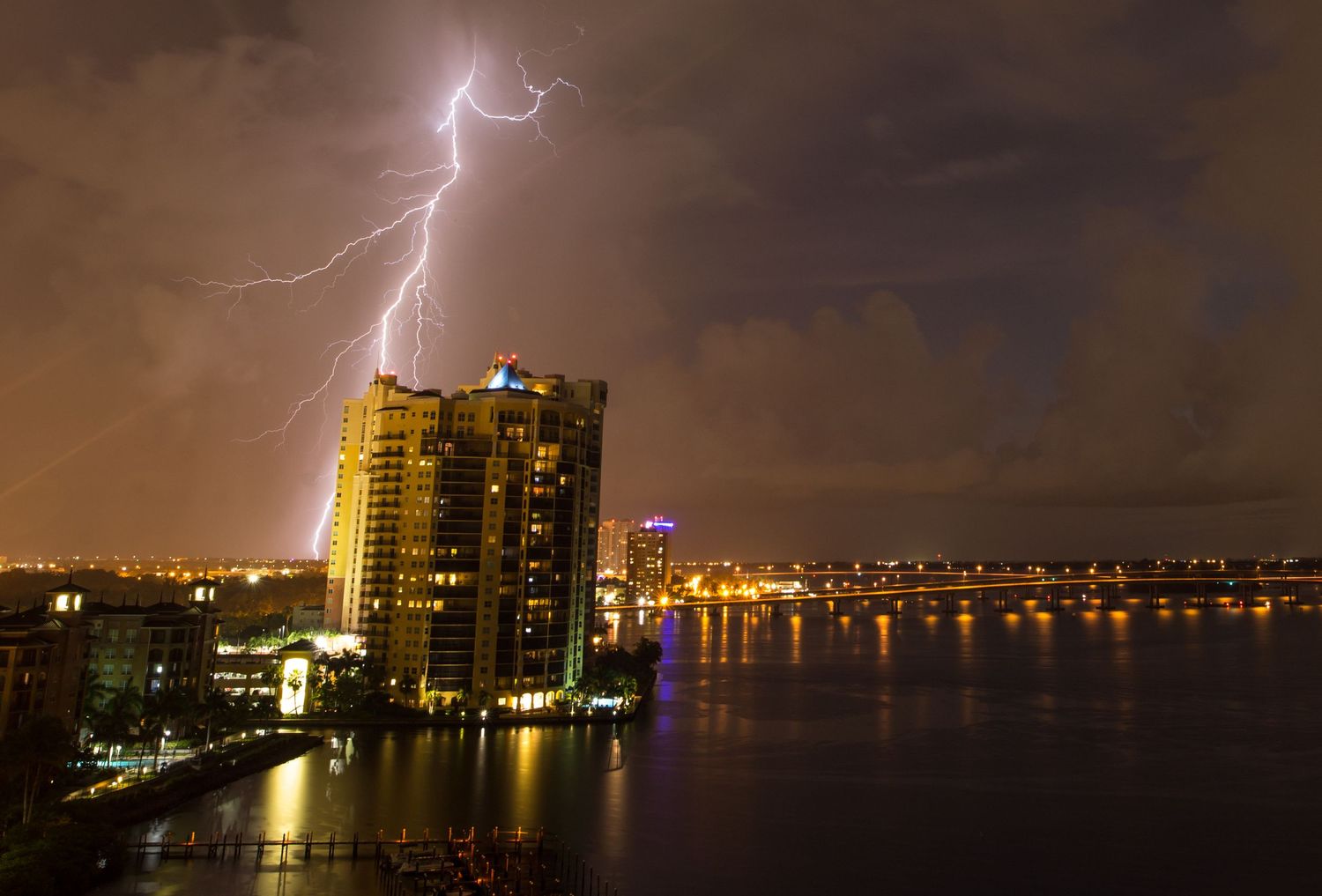 Lightning strikes illuminate the night sky over a city skyline with tall buildings reflected in calm waters.