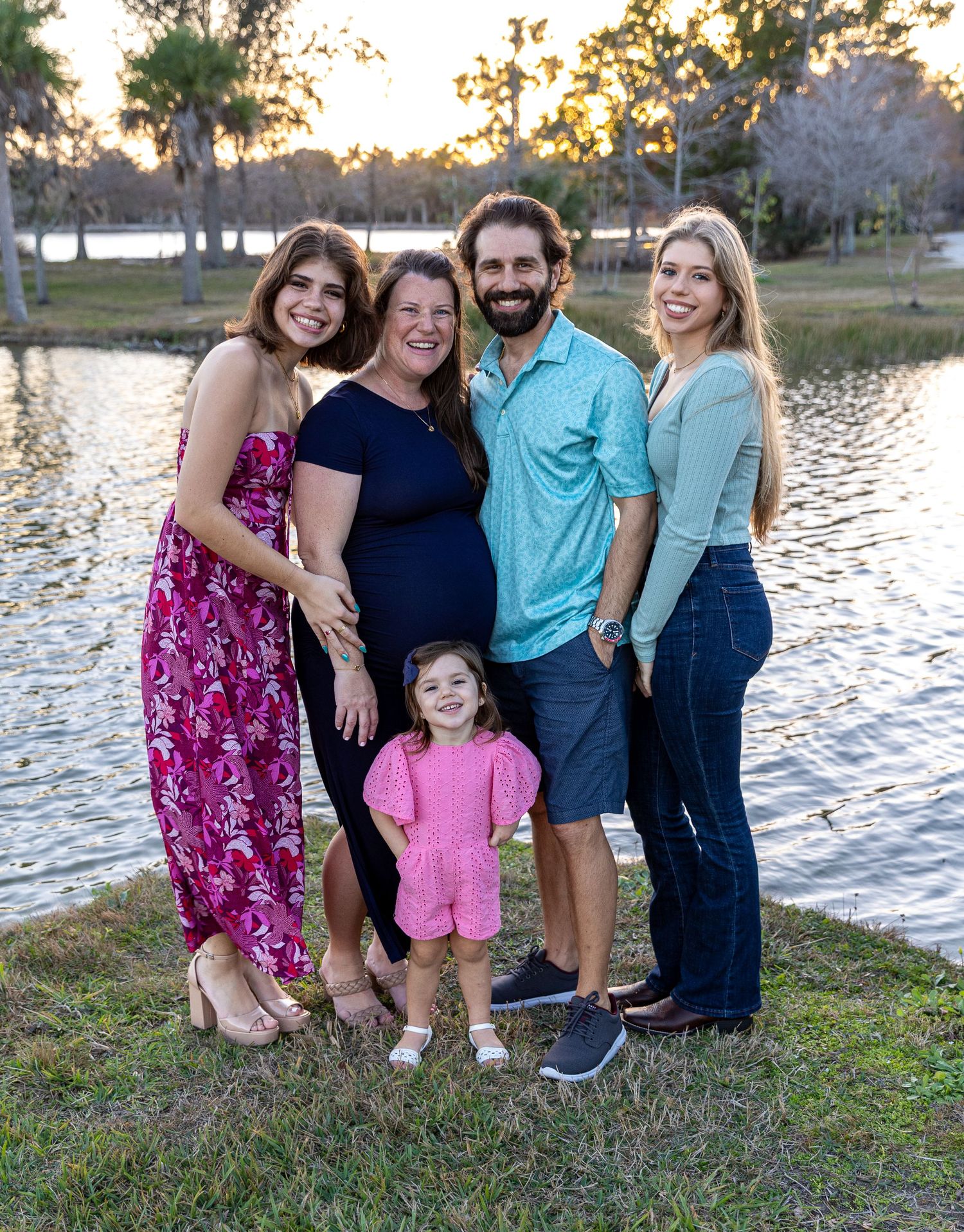 A family poses together by a lake at sunset, wearing casual clothes and smiling against the water backdrop.