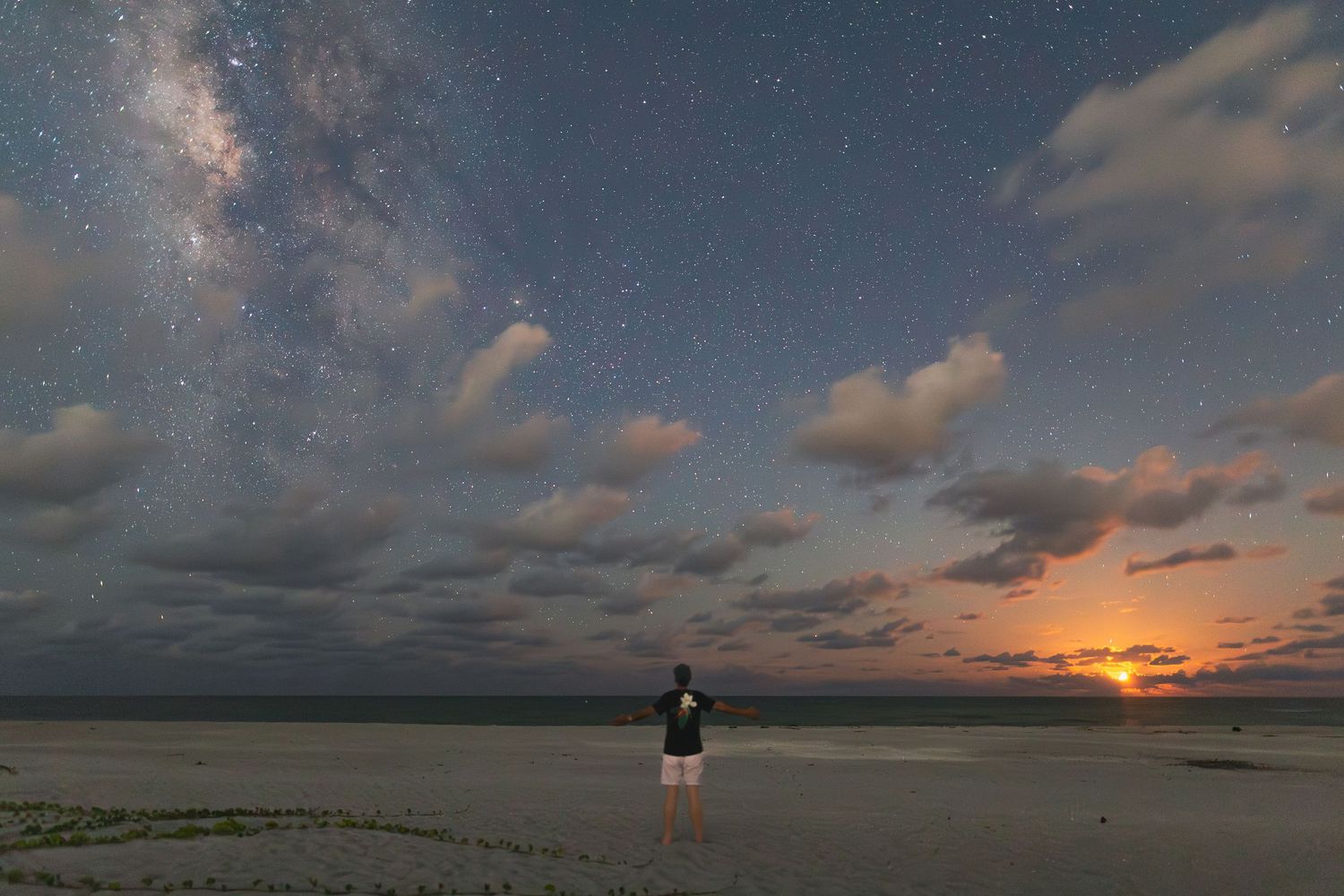 A silhouetted figure stands on a beach under a dramatic sunset sky with scattered clouds and visible stars.