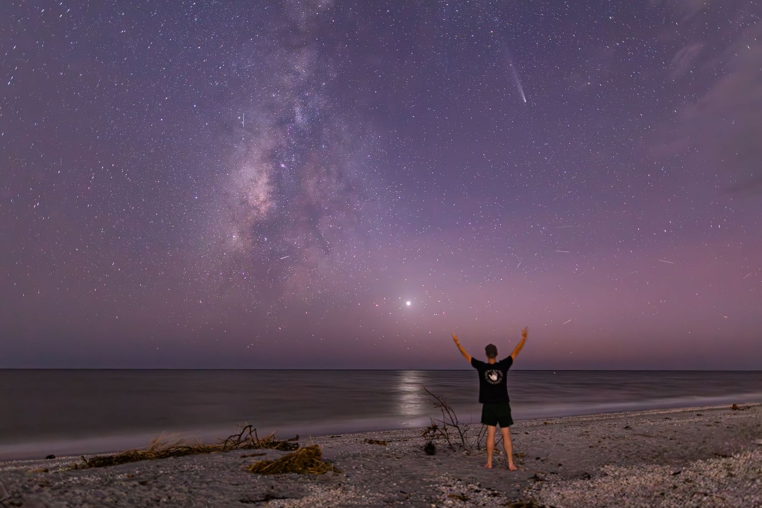 A silhouetted person stands on a beach with raised arms under the Milky Way galaxy and starlit purple night sky.
