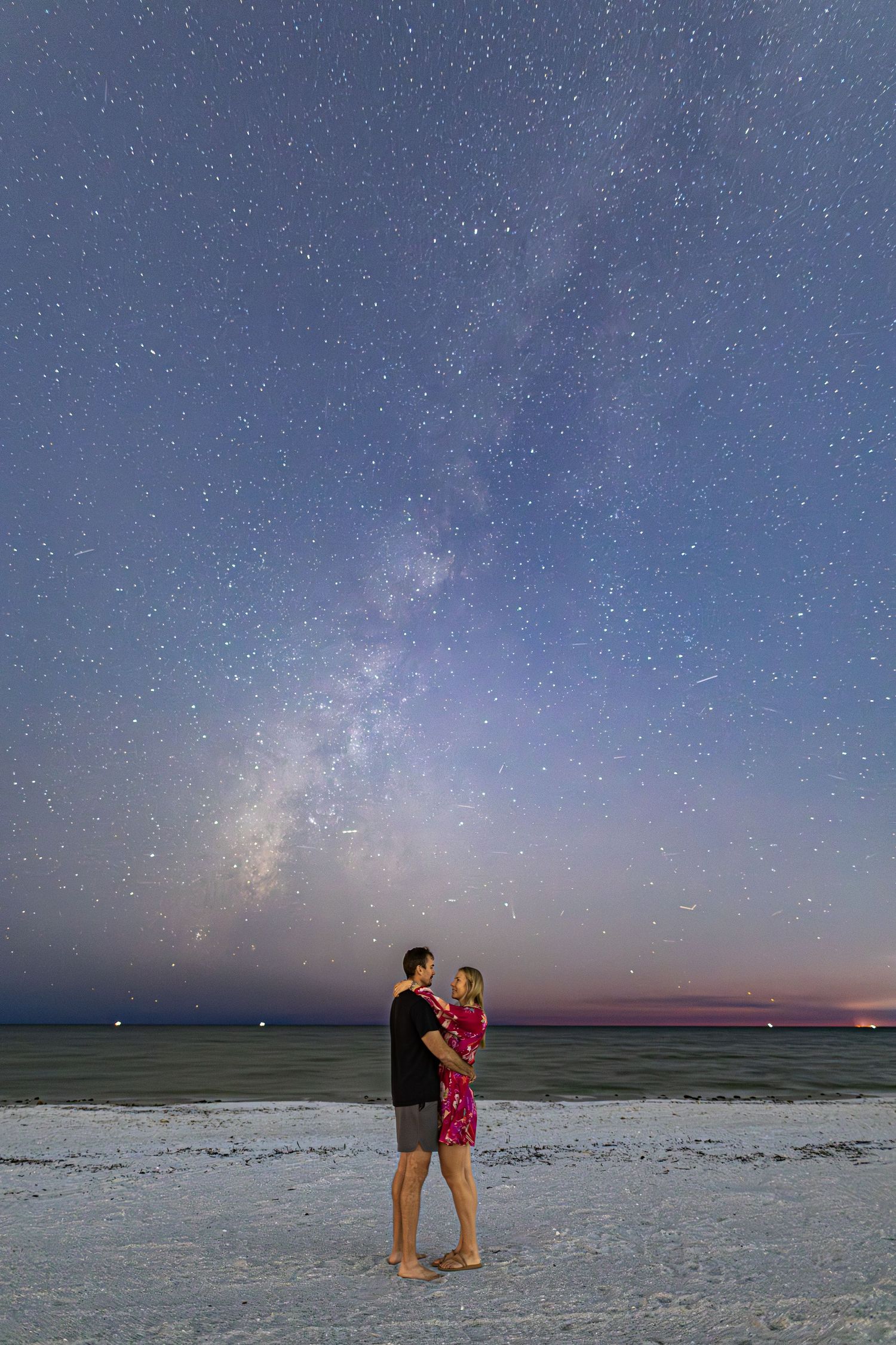 A couple embraces on a beach at night under a stunning Milky Way galaxy and starlit sky.