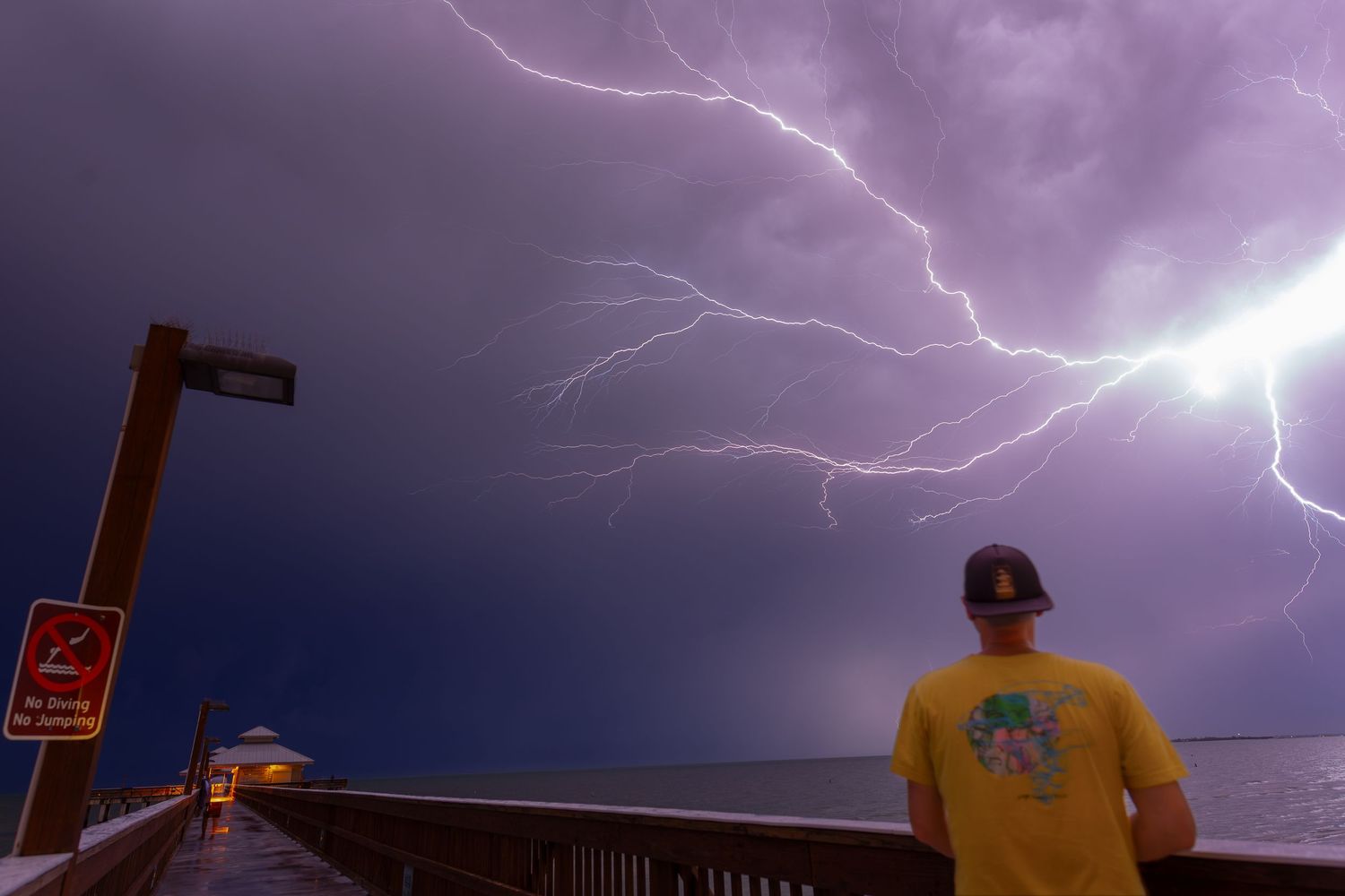 A person in a yellow shirt watches a dramatic lightning strike illuminate the stormy purple sky over a pier.
