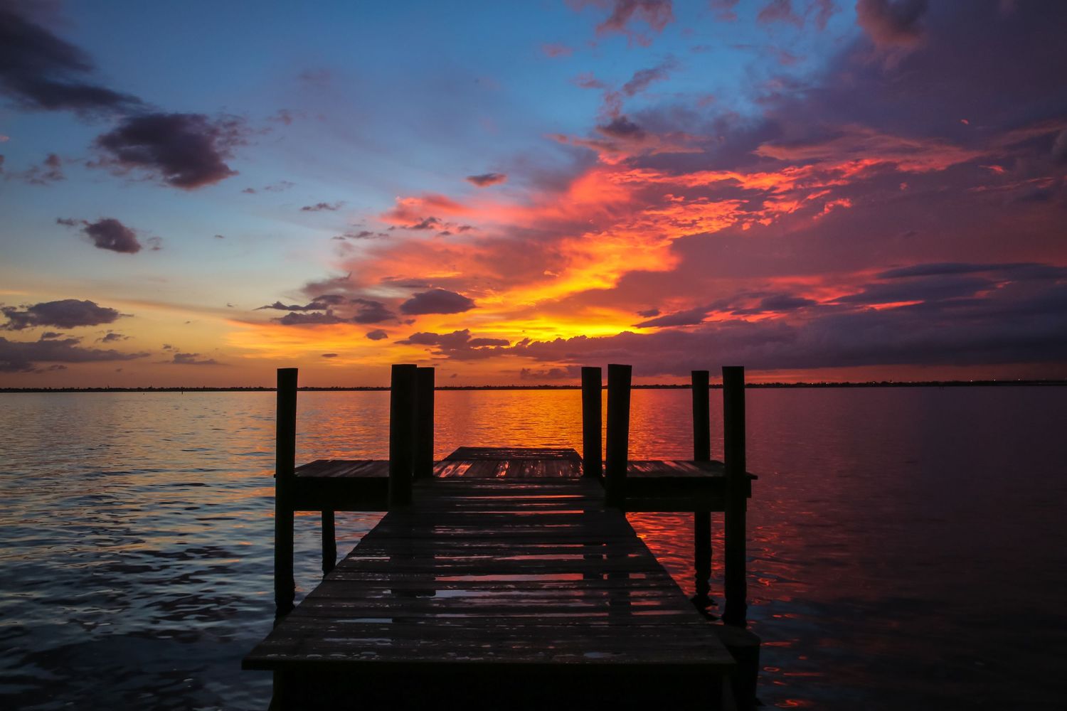 Wooden dock extends over calm water during dramatic sunset with vibrant orange and purple clouds reflected on surface.