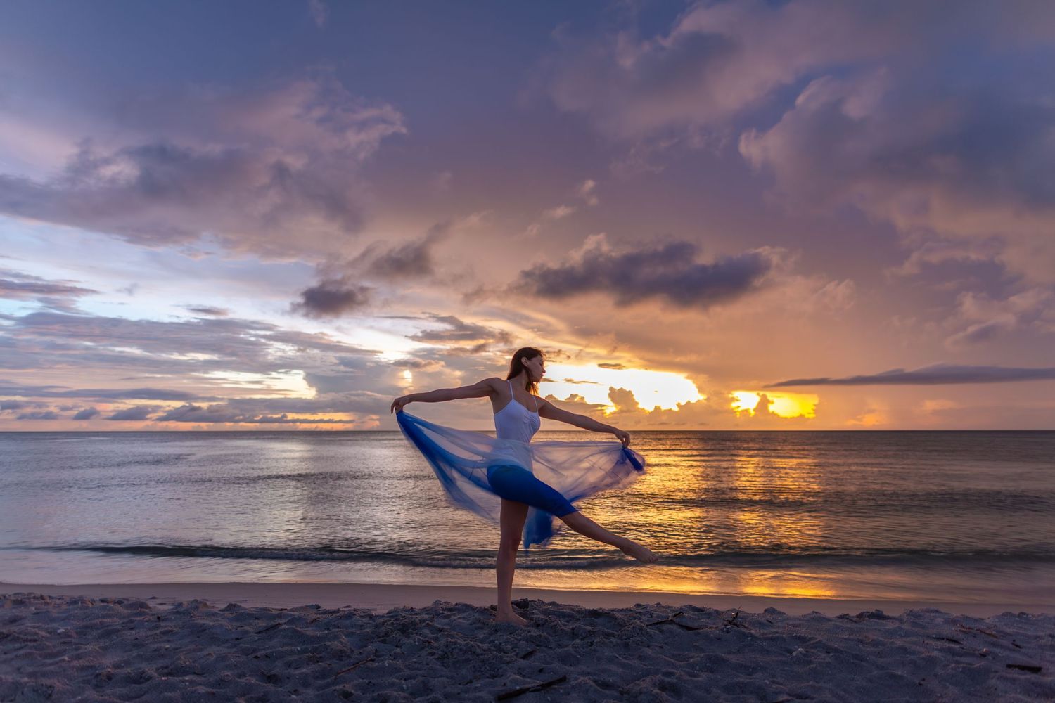 A silhouetted dancer strikes a graceful pose on the beach during a vibrant orange and purple sunset over the ocean.