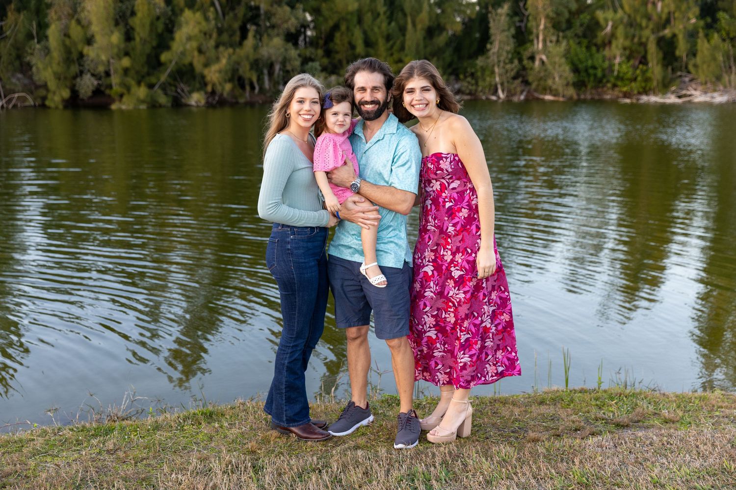 A family poses together on the grassy shore of a lake with trees reflecting in the calm water.