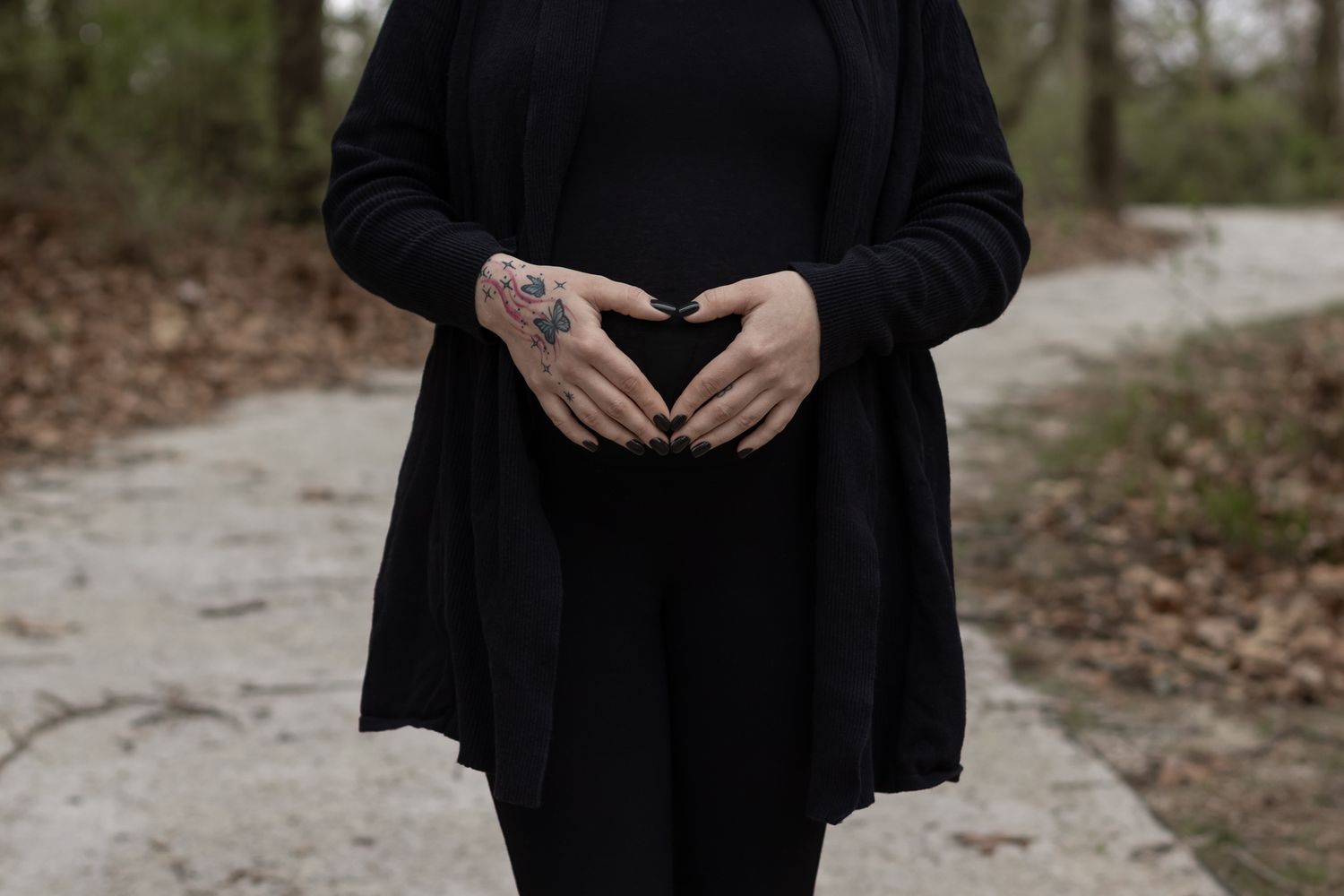 Hands form a heart shape over a black dress while standing on a dirt path in moody lighting.