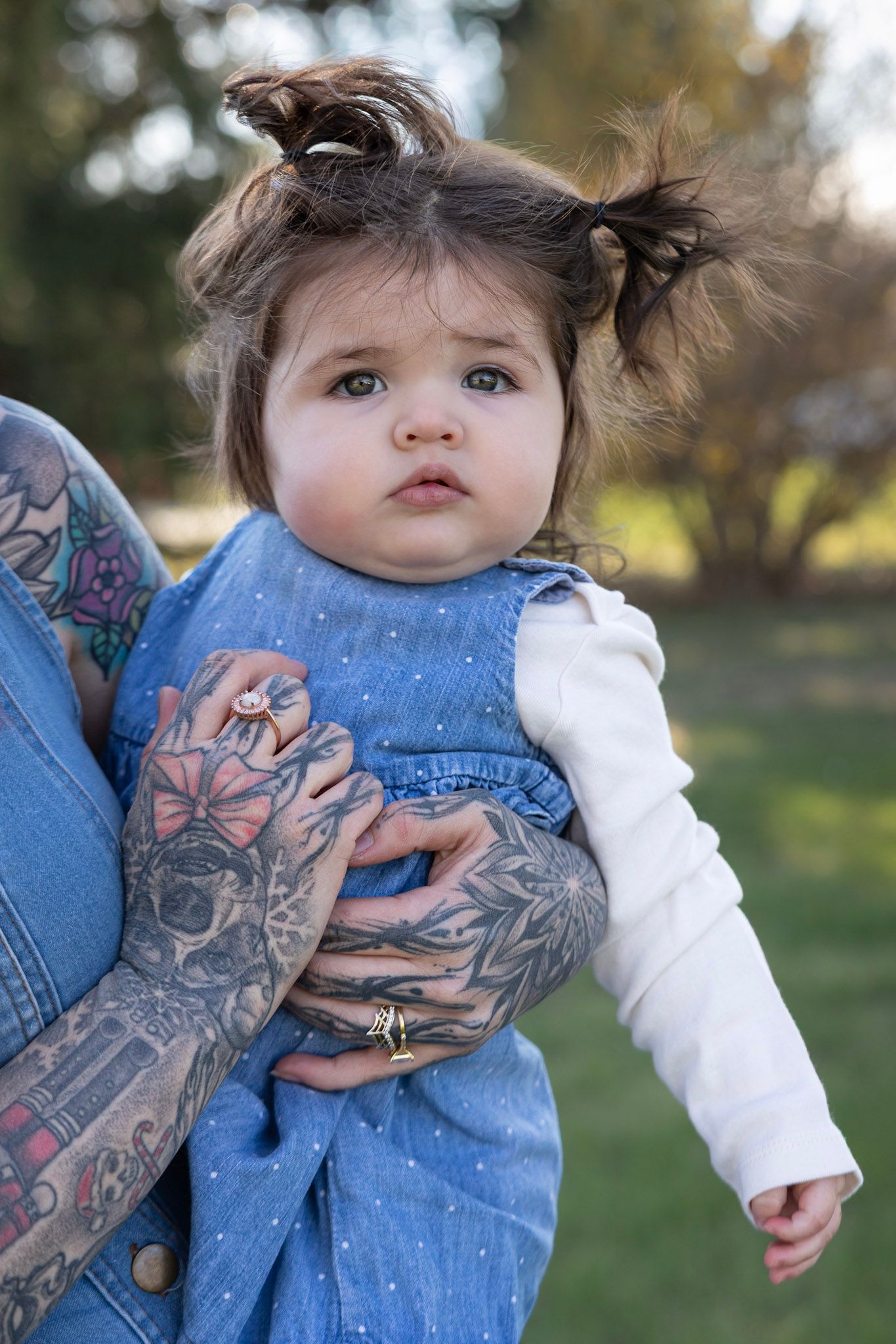 A small child in a blue denim dress being held by tattooed arms outdoors in natural lighting.