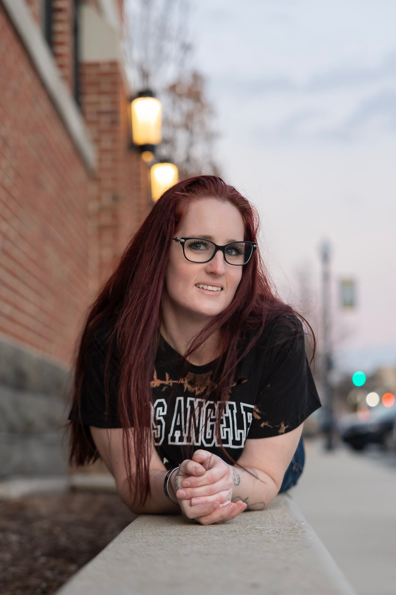 Woman with long red hair wearing glasses and a Los Angeles t-shirt leans on a brick wall at dusk.