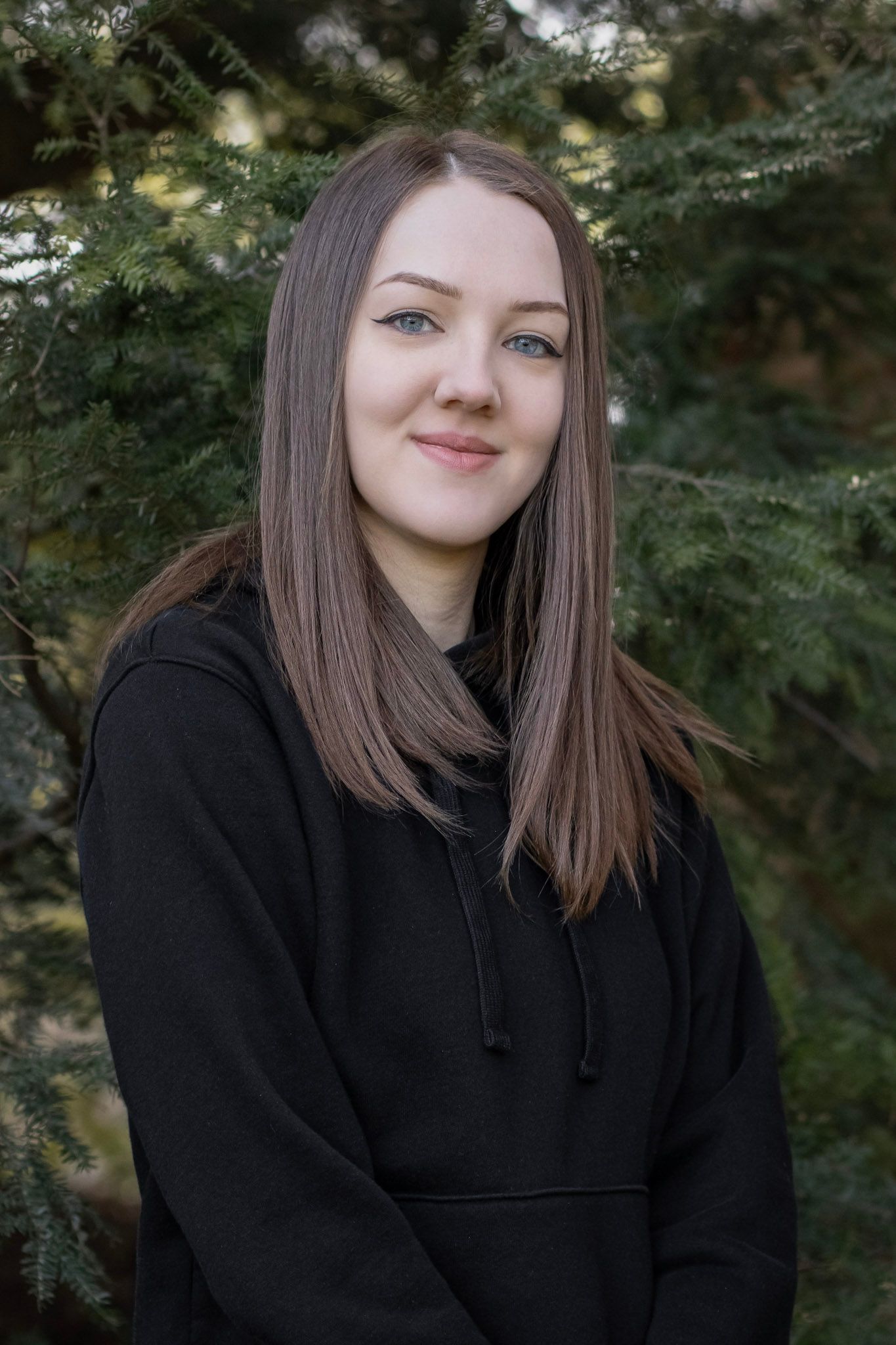 Portrait of a woman wearing a black hoodie outdoors with evergreen trees in the background.