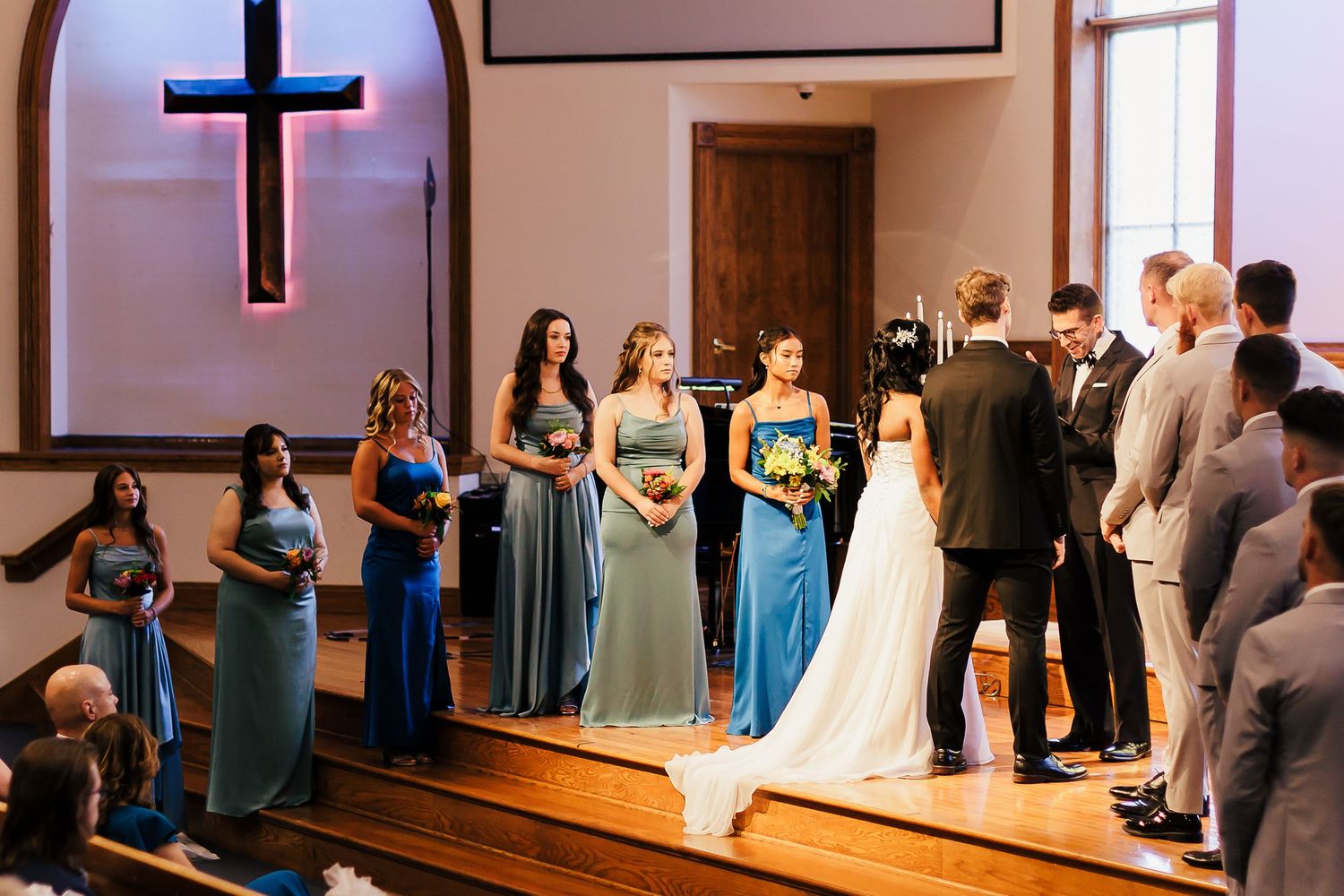 Church wedding ceremony with bridesmaids in blue dresses standing at altar under illuminated cross.