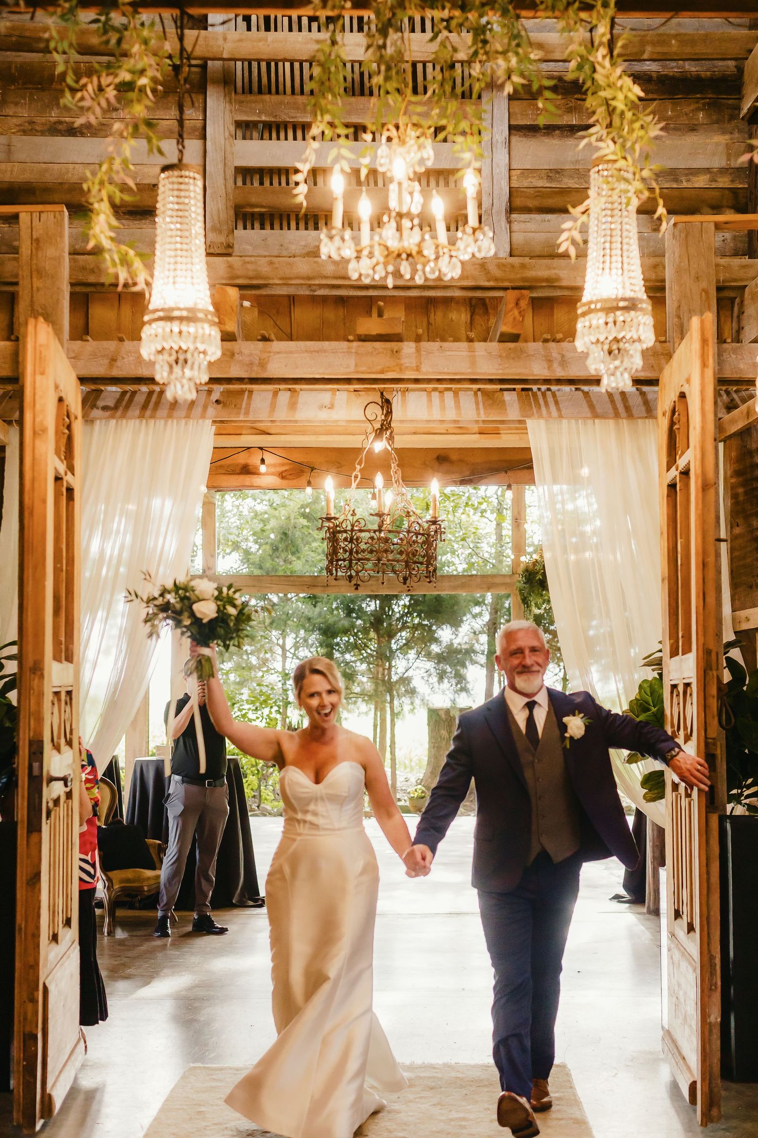 Newlyweds hold hands and dance joyfully in a rustic barn venue decorated with elegant crystal chandeliers.