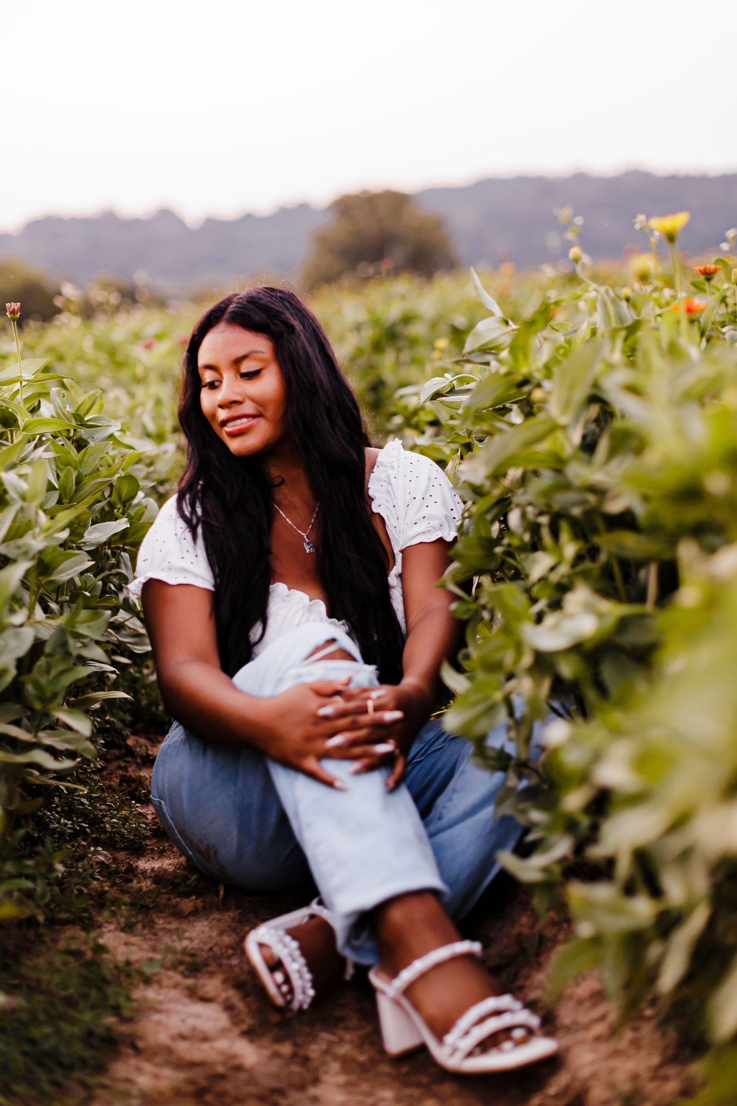 Person in white top and jeans sitting casually in a field during golden hour.