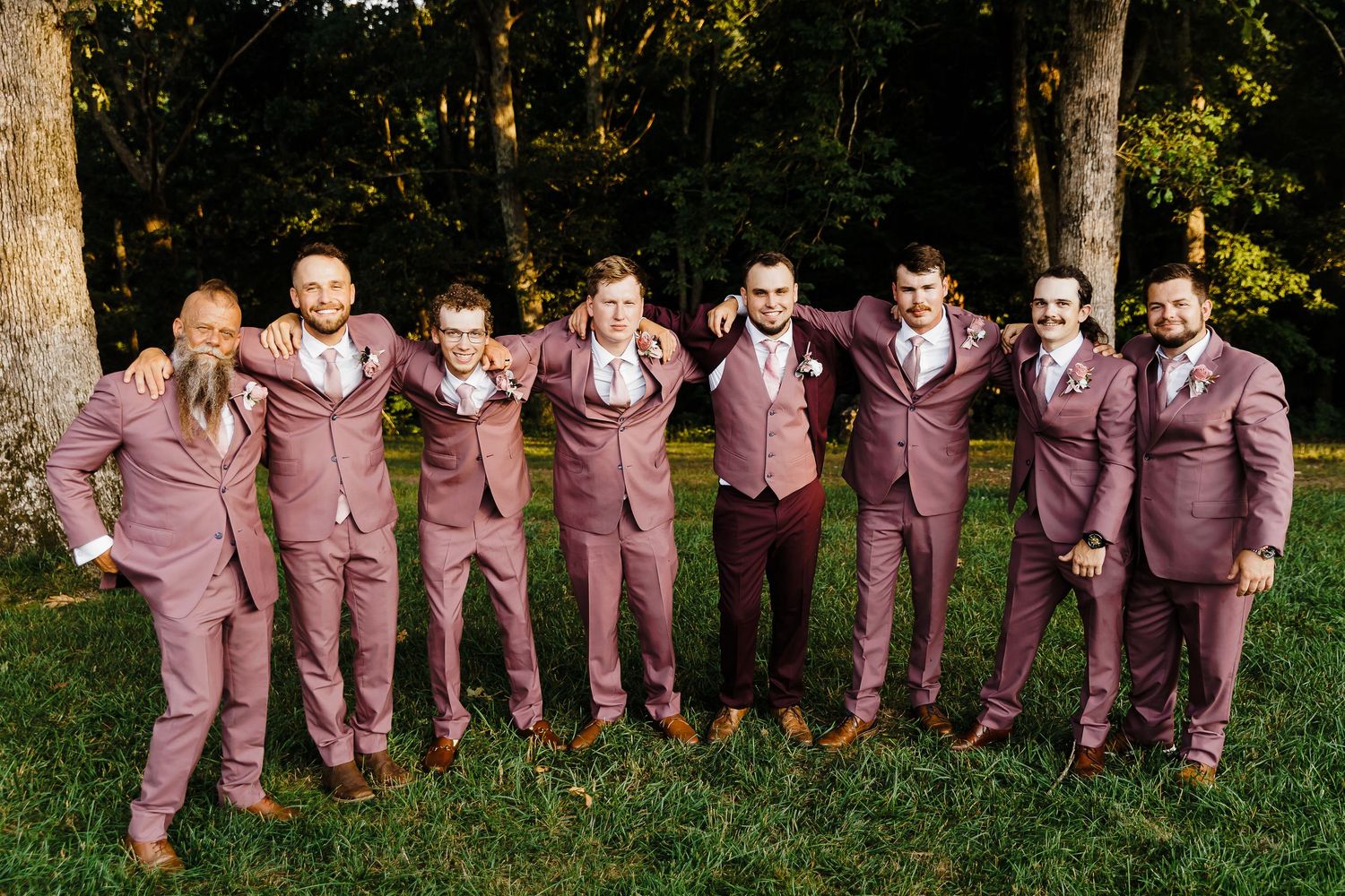 Groomsmen in matching mauve suits pose together on grass with trees in background.