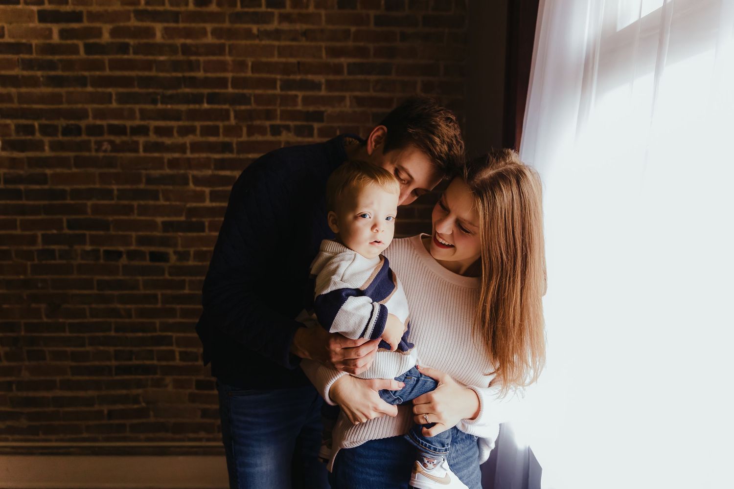 Indoor family photoshoot with natural window light against brick wall background.
