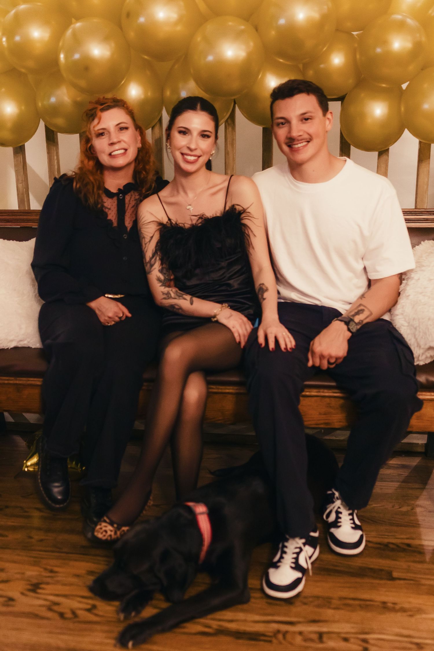 A festive gathering with gold balloons decorating the background and a black dog sitting at their feet.