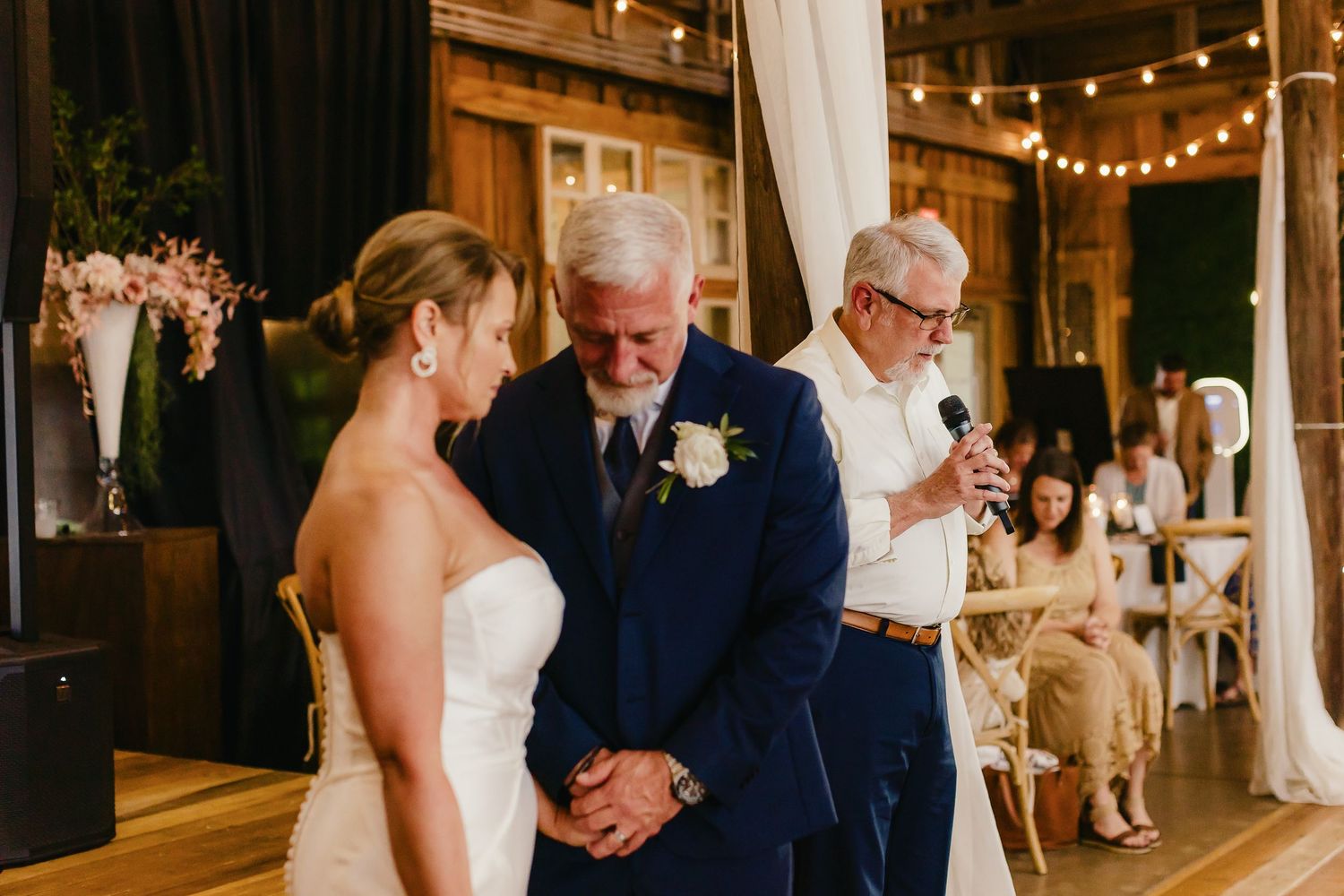 A couple shares a romantic dance at their wedding reception under warm string lights and white drapes.