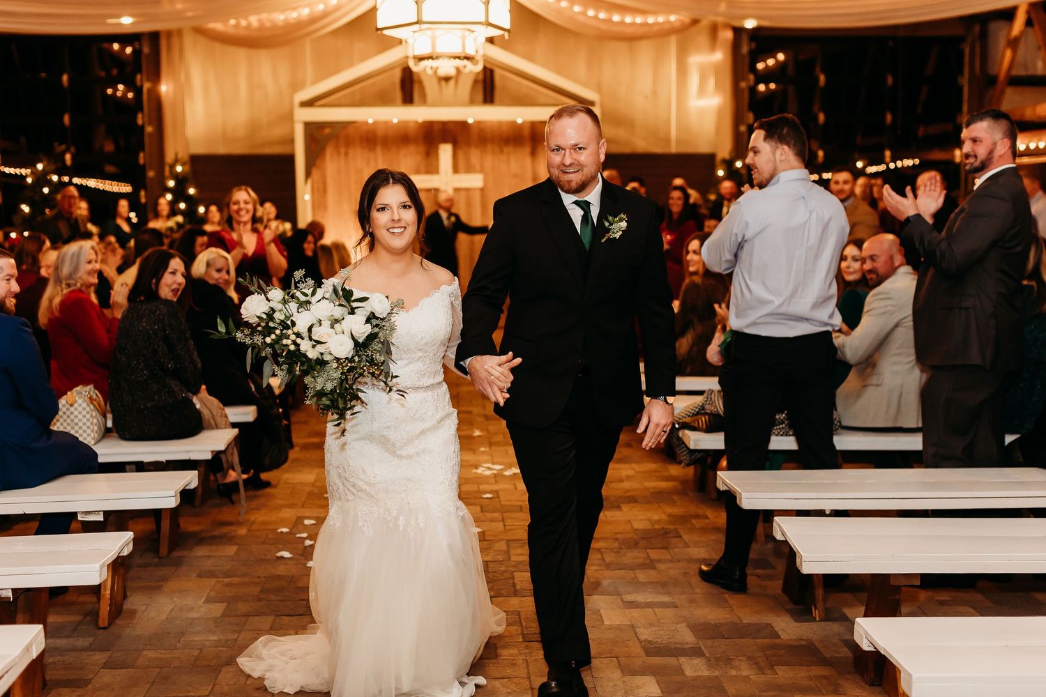 Wedding ceremony recessional in warmly lit venue with wooden benches and white decor.