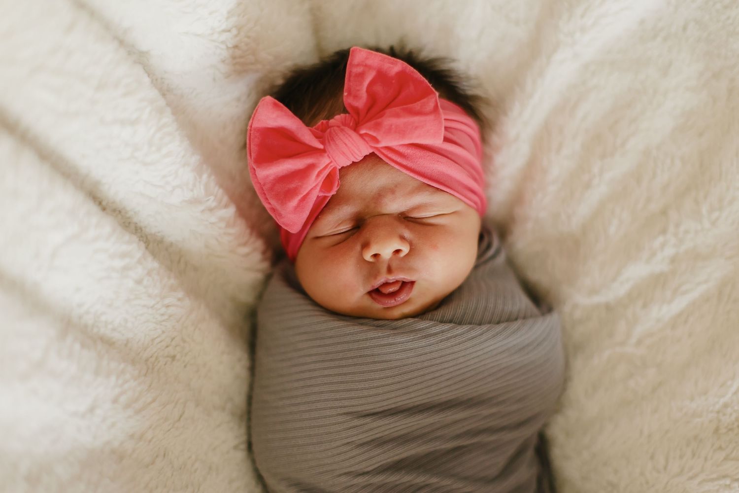 Peaceful sleeping baby wrapped in gray swaddle with pink bow headband.