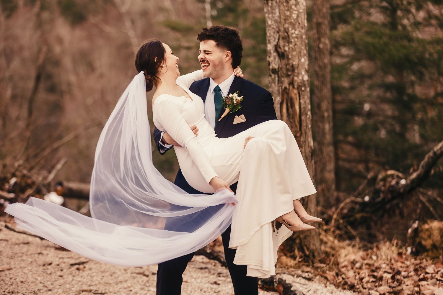 A romantic wedding couple embraces in a wooded forest setting as a long white veil flows in the breeze.