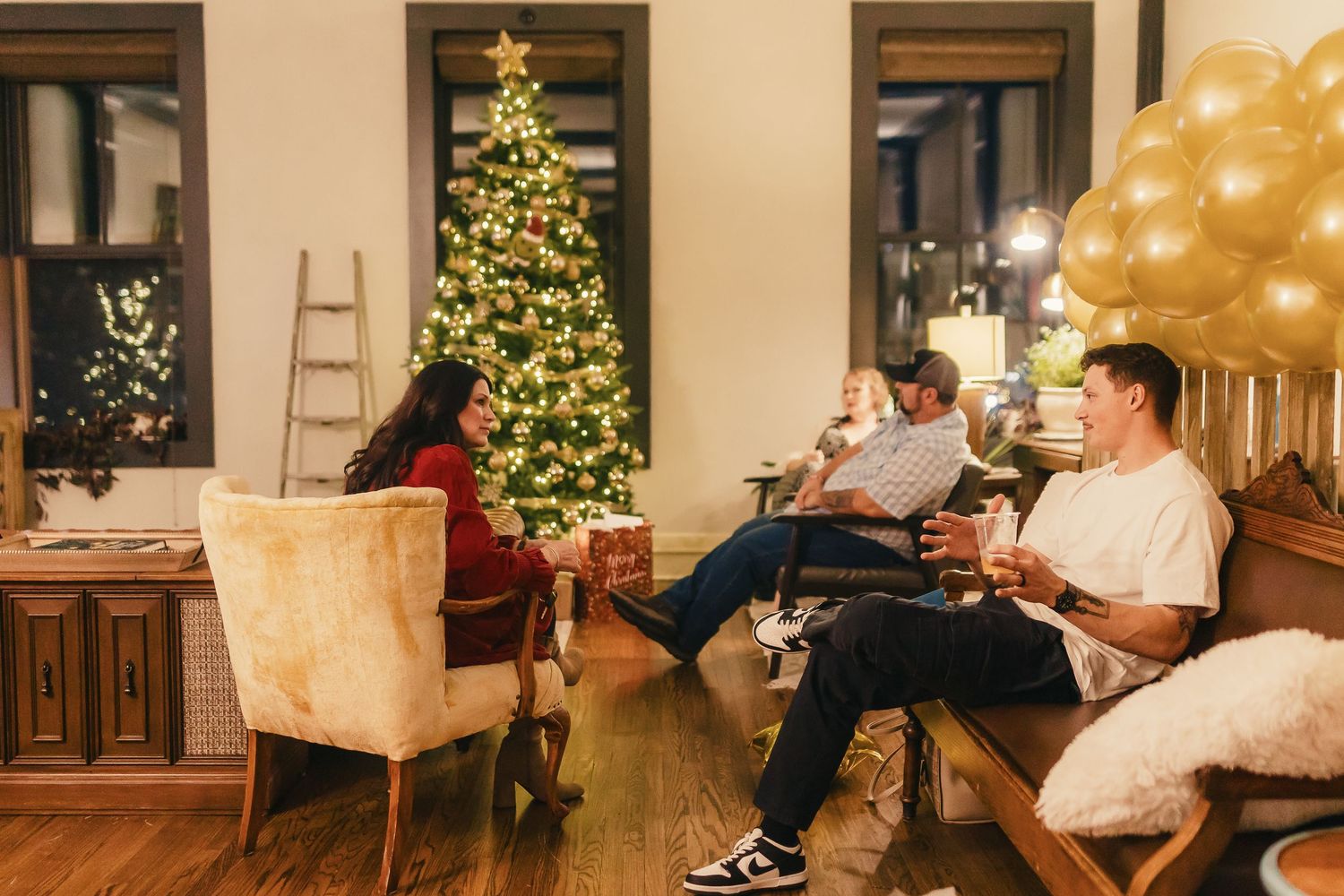 Friends relaxing in a cozy living room decorated for Christmas with a lit tree and golden balloons in the background.
