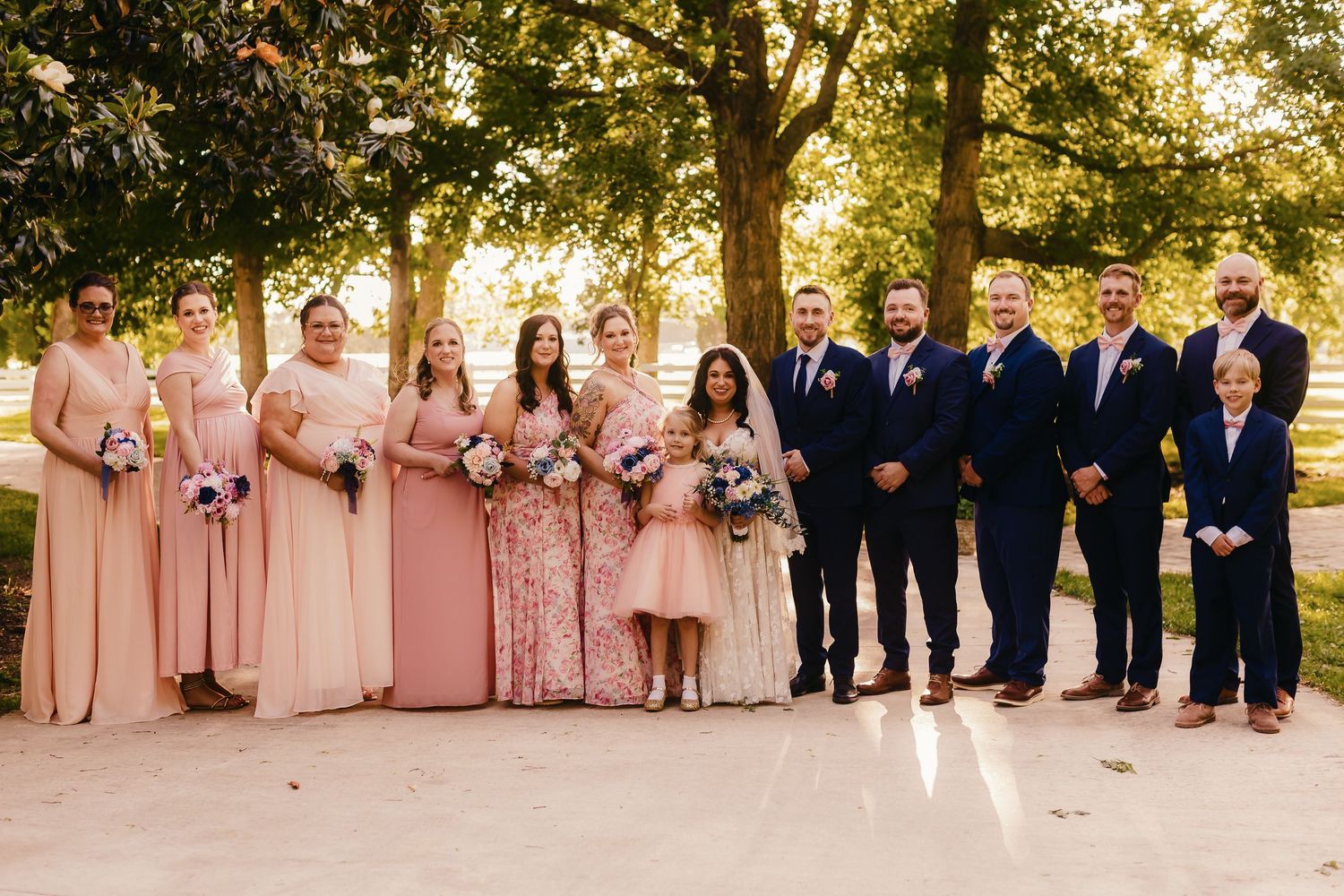 Wedding party posing outdoors, bridesmaids in blush pink gowns and groomsmen in navy suits, with bride and groom centered.