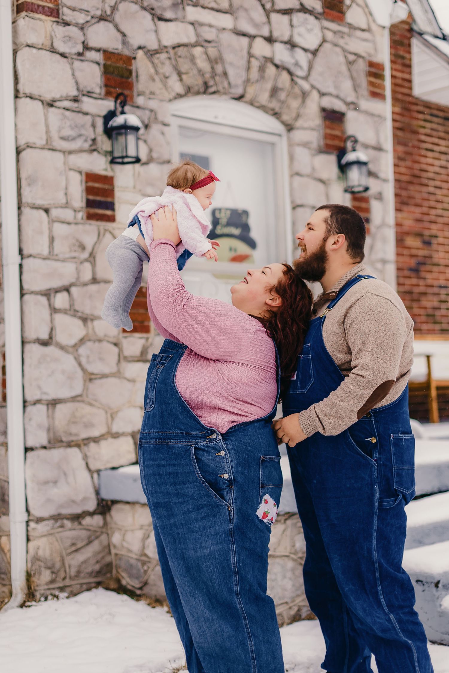Family wearing matching denim overalls plays together in the snow against a stone wall.